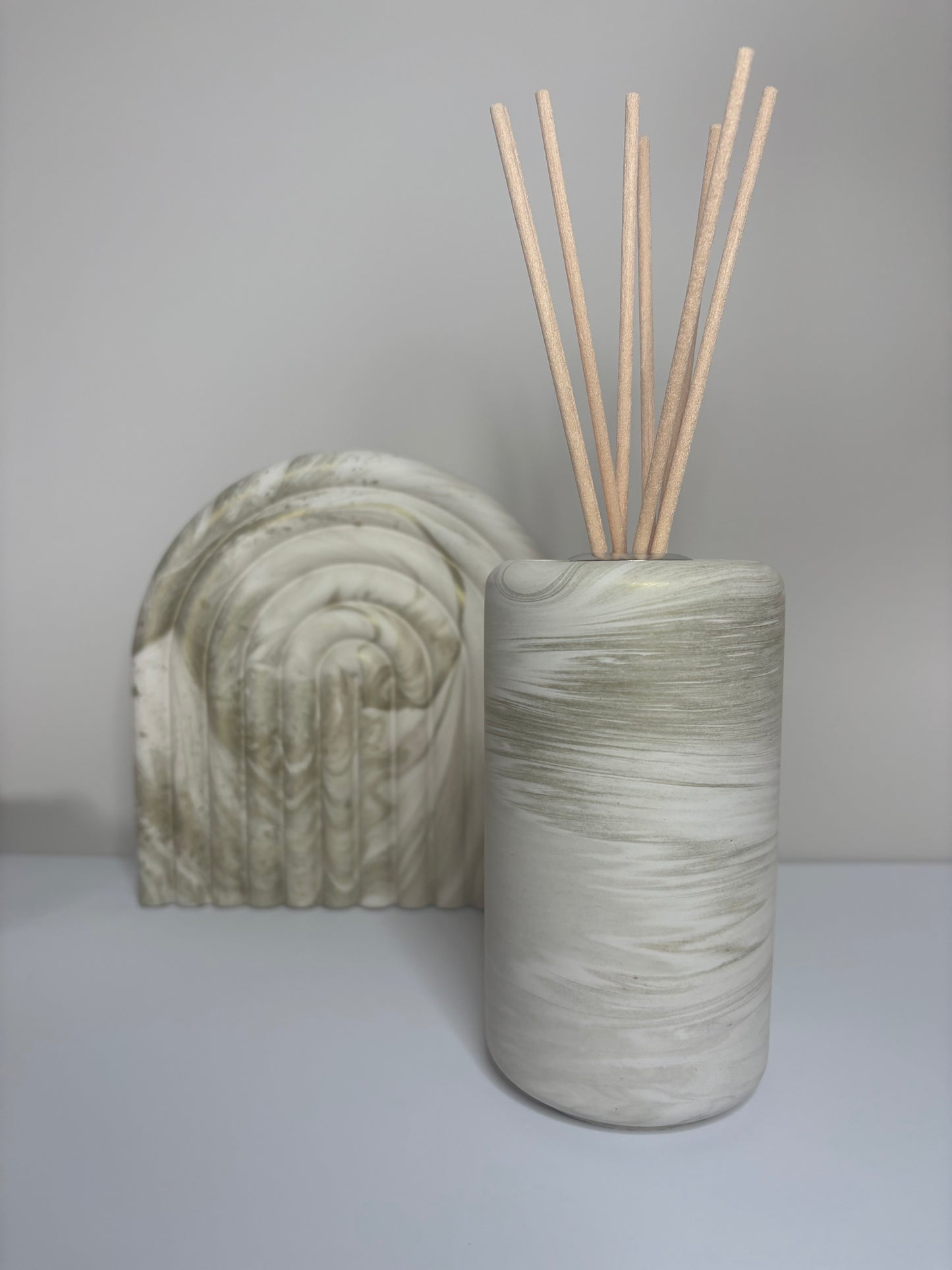 Handmade Solace Diffuser in a tall cylindrical Jesmonite vessel with soft marbled beige and stone grey tones, styled beside a matching Luna Arch Bookend on a minimalist surface for a cohesive, neutral display.
