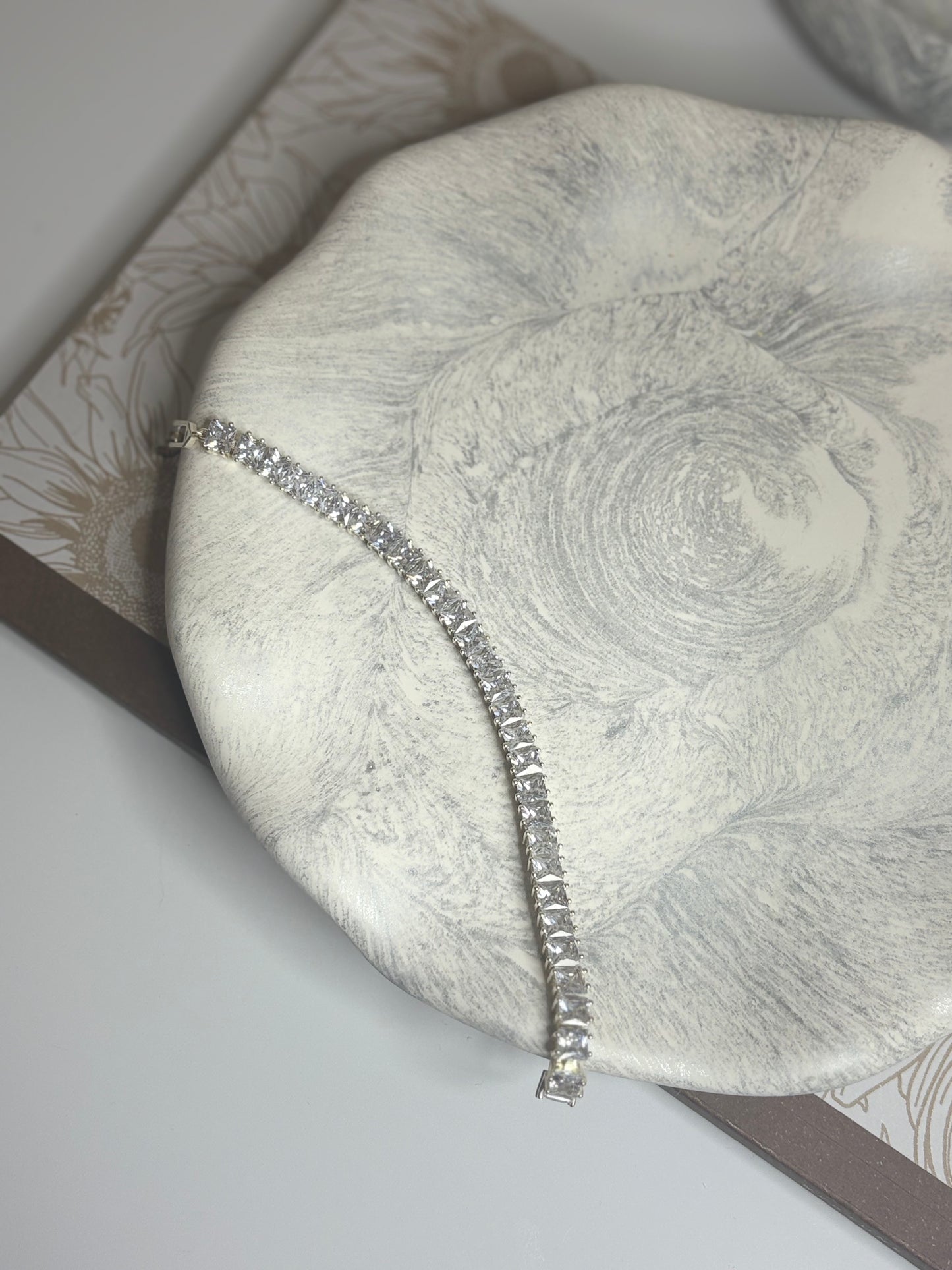 A handmade round decorative plate with a soft grey and white marbled design, featuring a silver tennis bracelet laid elegantly across its surface. The plate rests on a floral-patterned book, showcasing its smooth matte texture and wavy organic edge.