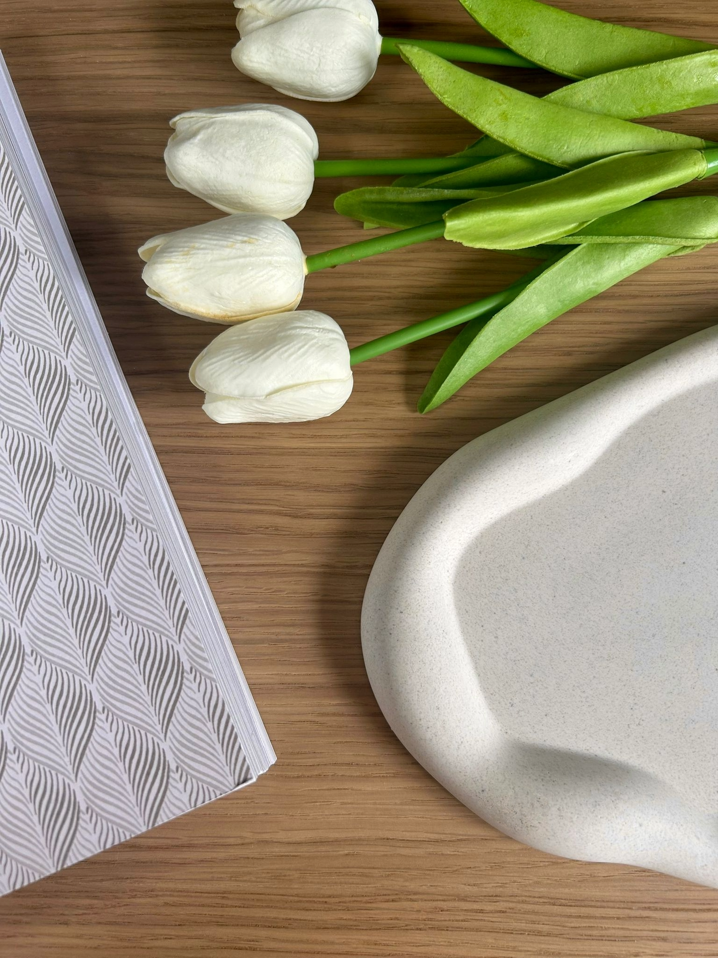 Flatlay of a modern ceramic home decor tray paired with faux white tulips and a patterned journal, showcasing handmade minimalist design aesthetics.