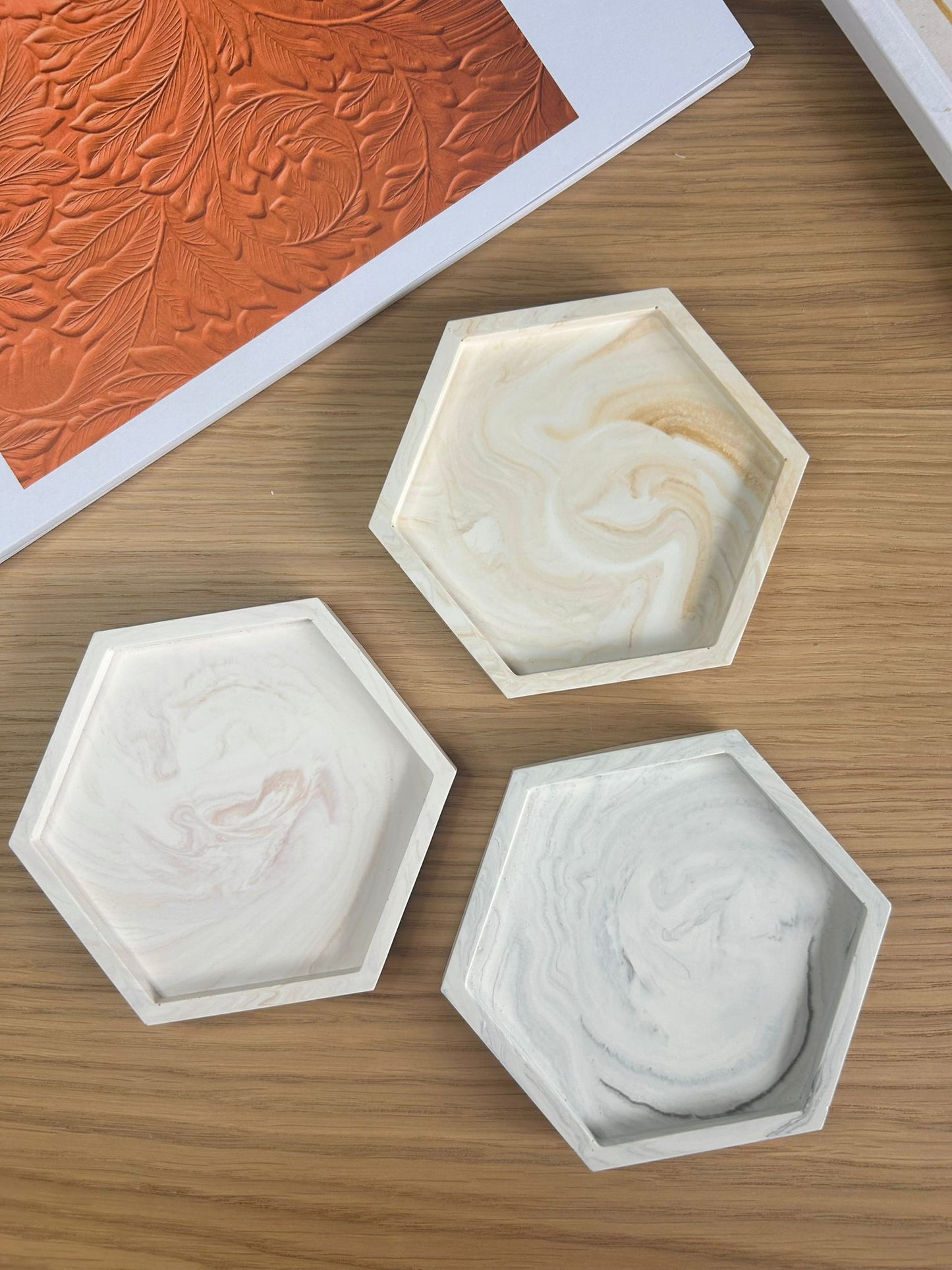 Set of three hexagonal handmade Jesmonite trays with marble finishes in warm beige, pink and grey. Sustainable, minimalist home accessories styled on a wooden surface.