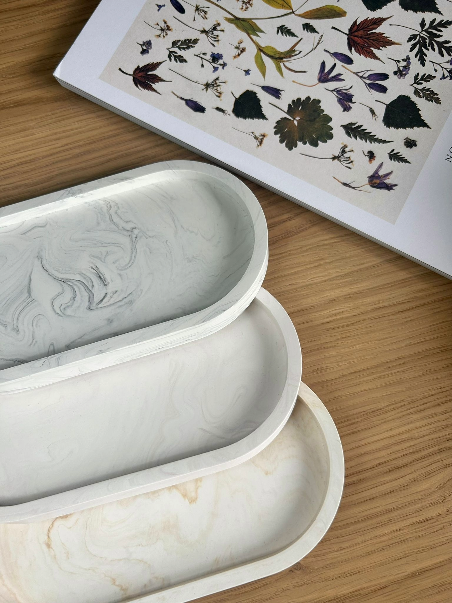 Set of three handmade Jesmonite trays in grey, ivory, and warm cream marble finishes, styled beside a botanical book – sustainable and versatile home accessories.