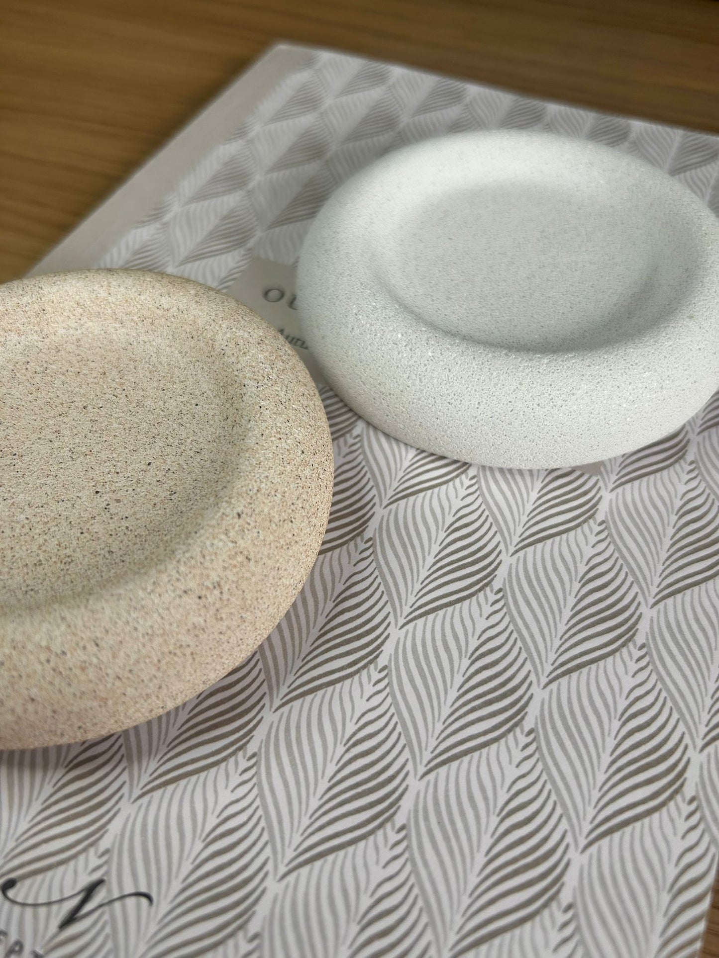 Two round Jesmonite trays with natural speckled textures in beige and white, displayed on a patterned book. A sculptural set of handmade home accessories.