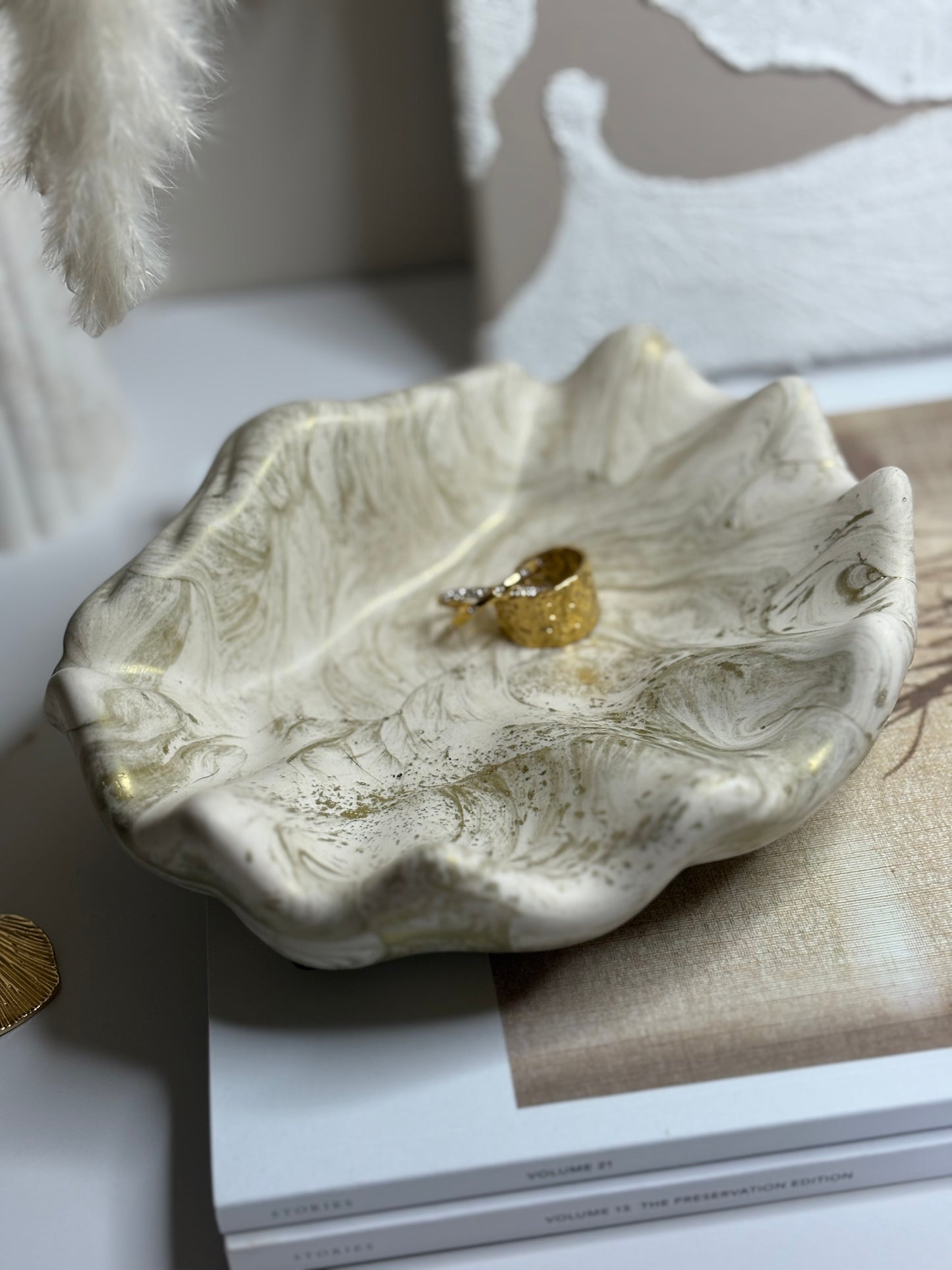 A handmade Jesmonite shell-shaped trinket dish with soft ivory and gold marbling, displayed on stacked neutral-toned books. The dish holds a single gold textured ring, showcasing its organic rippled edges and elegant, sculptural design within a calm, minimalist setting.