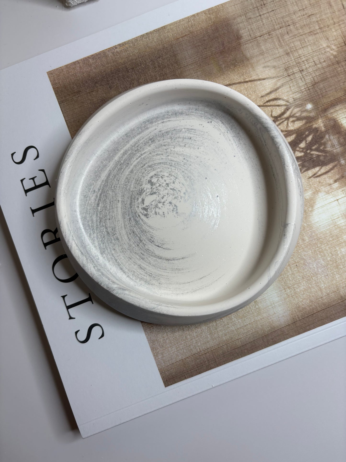 A handmade Jesmonite trinket dish with a soft grey and white marbled swirl design, displayed on an open book with neutral tones in a minimalist setting.
