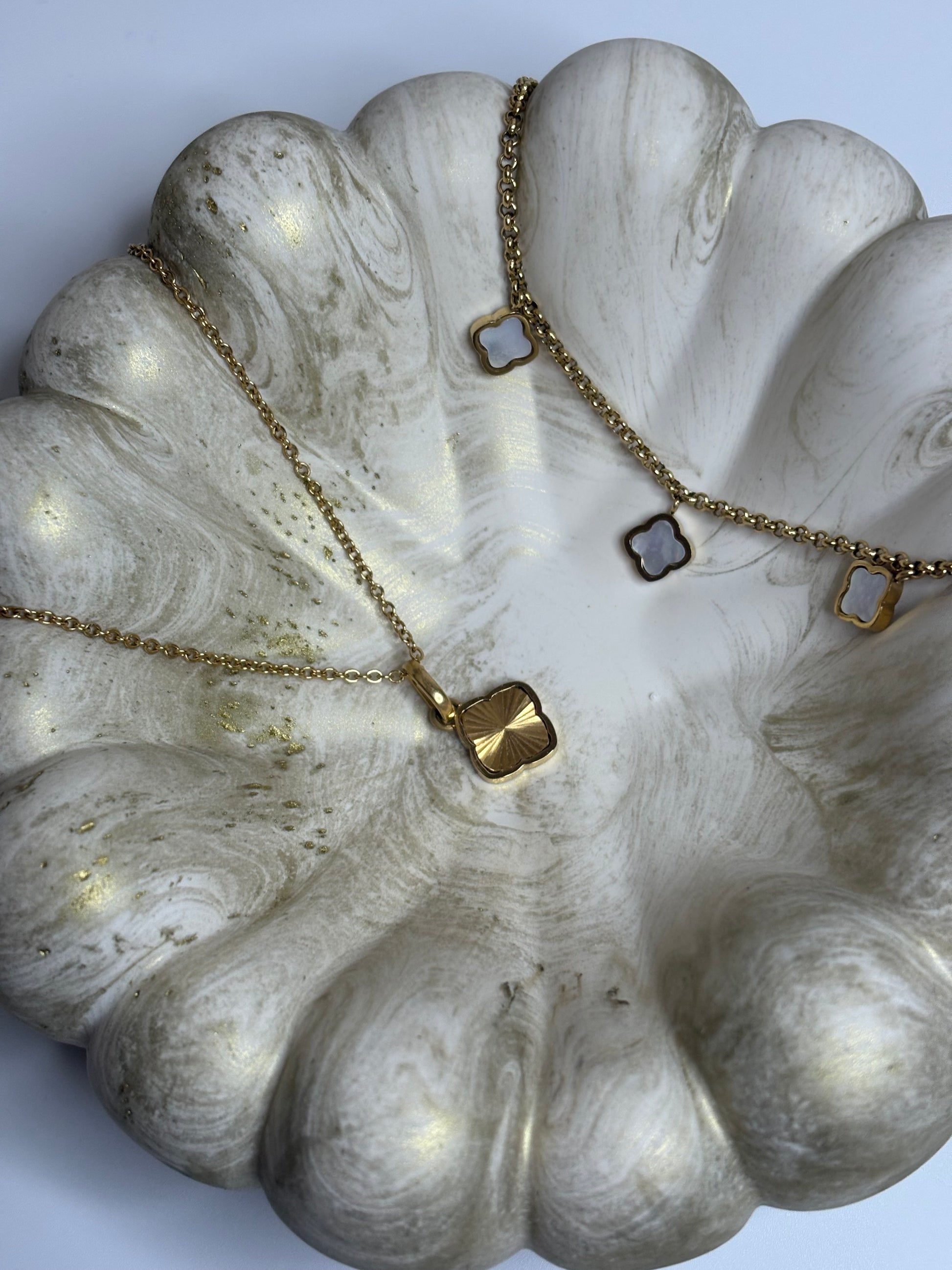 A handmade Jesmonite Celeste Trinket Bowl with a rippled, sculptural edge and soft gold marbled detailing, holding a delicate gold necklace with clover-shaped charms. The bowl’s smooth matte finish and organic form create an elegant, minimalist display on a neutral background.