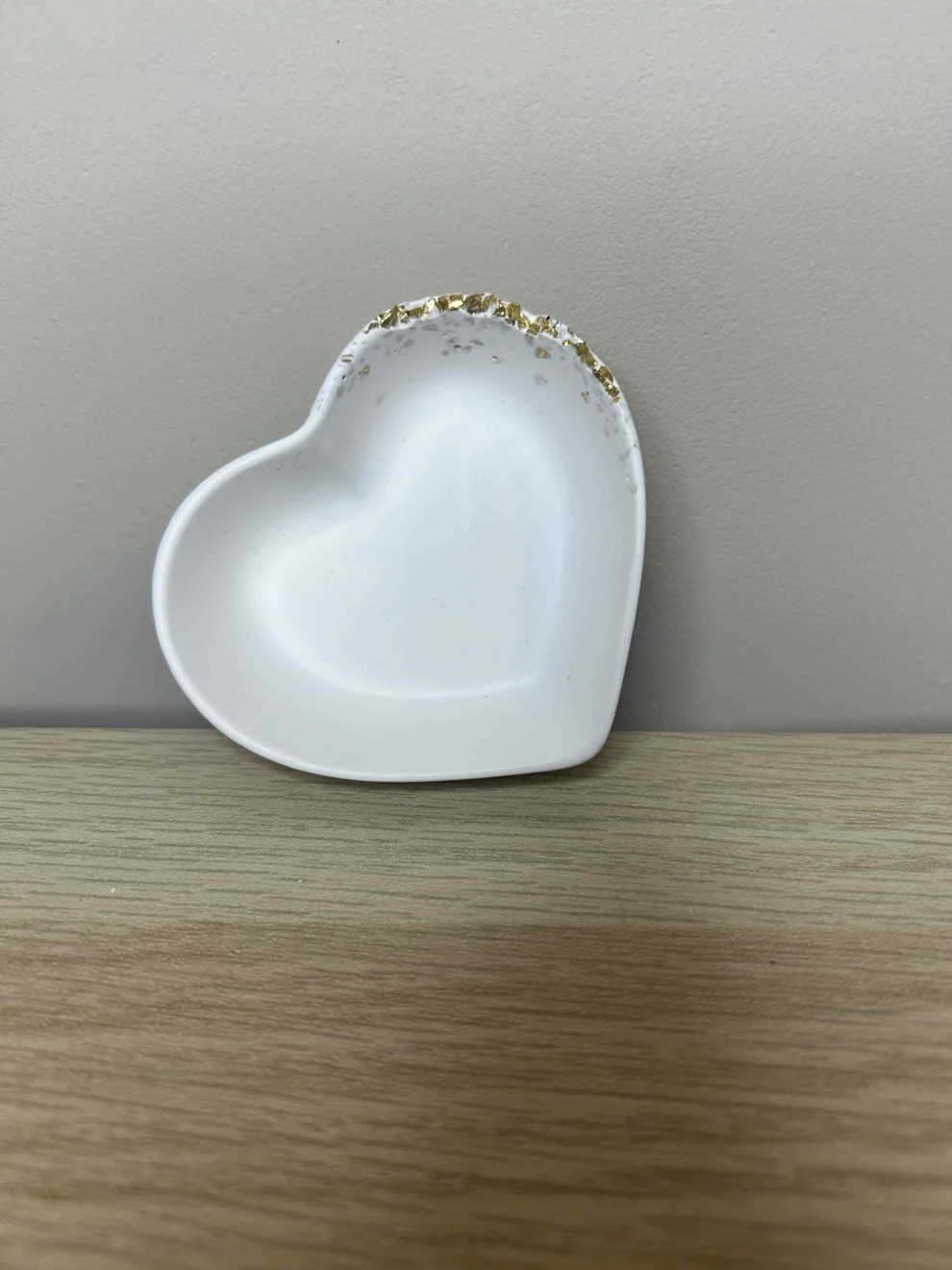 Handmade Home Accessories - a shot of the full small heart trinket dish in white with a raw gold druzy edge on the top right hand corner. The dish is leaning against a brown wall, with a wooden shelf underneath