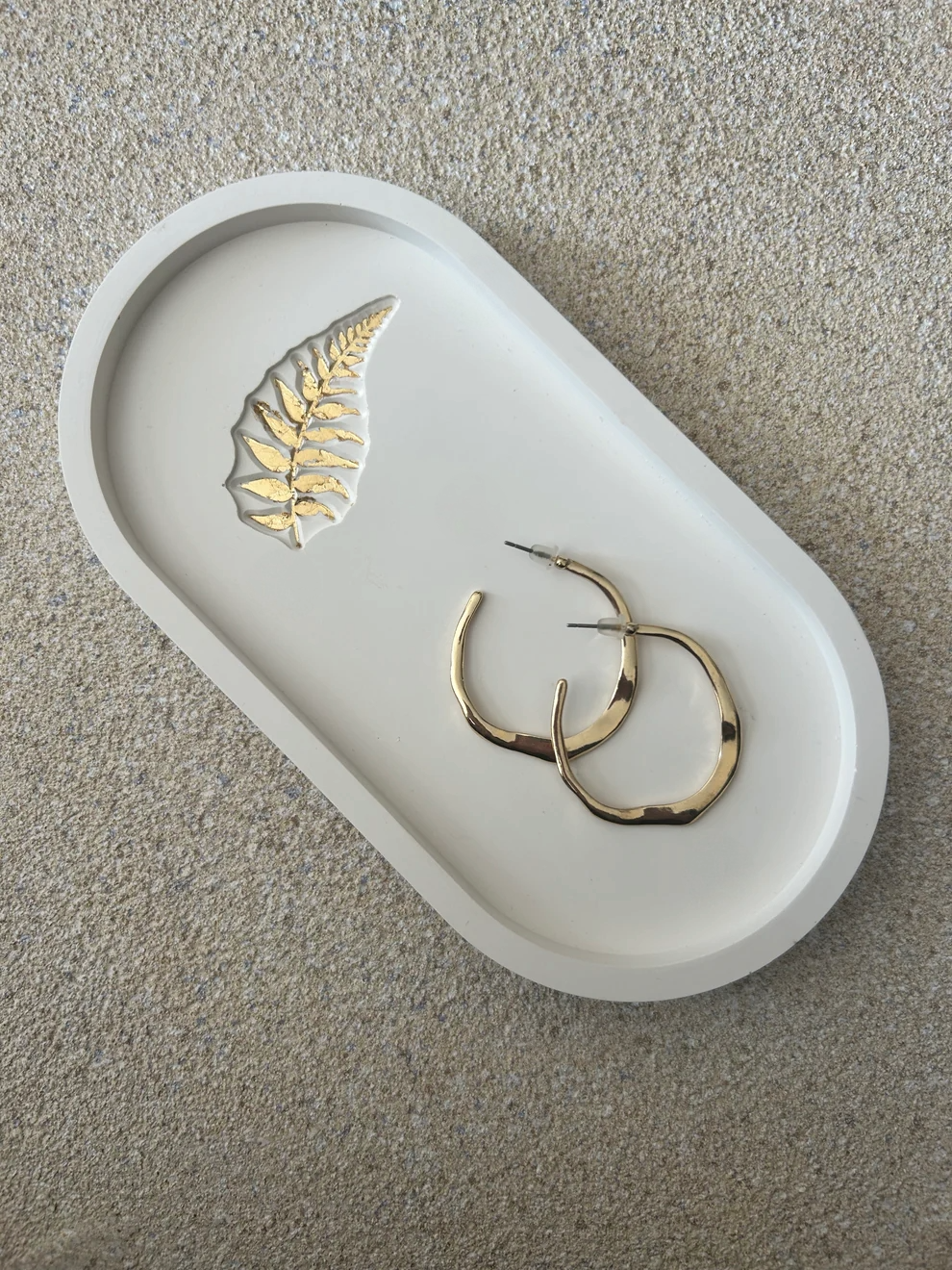 Handmade Home Accessories - A white oval tray at a 45 degree angle, with a gold leaf fern inlay. it has a pair of gold hoops displayed on it for decortive purposes on a natural stone backdrop.