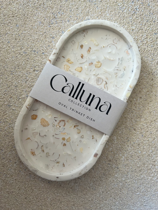 Handmade Home Accessories - A front view of a oval trinket tray that has a crushed seashell terrazzo design. it has a belly band around the centre with the company name Calluna Collection.