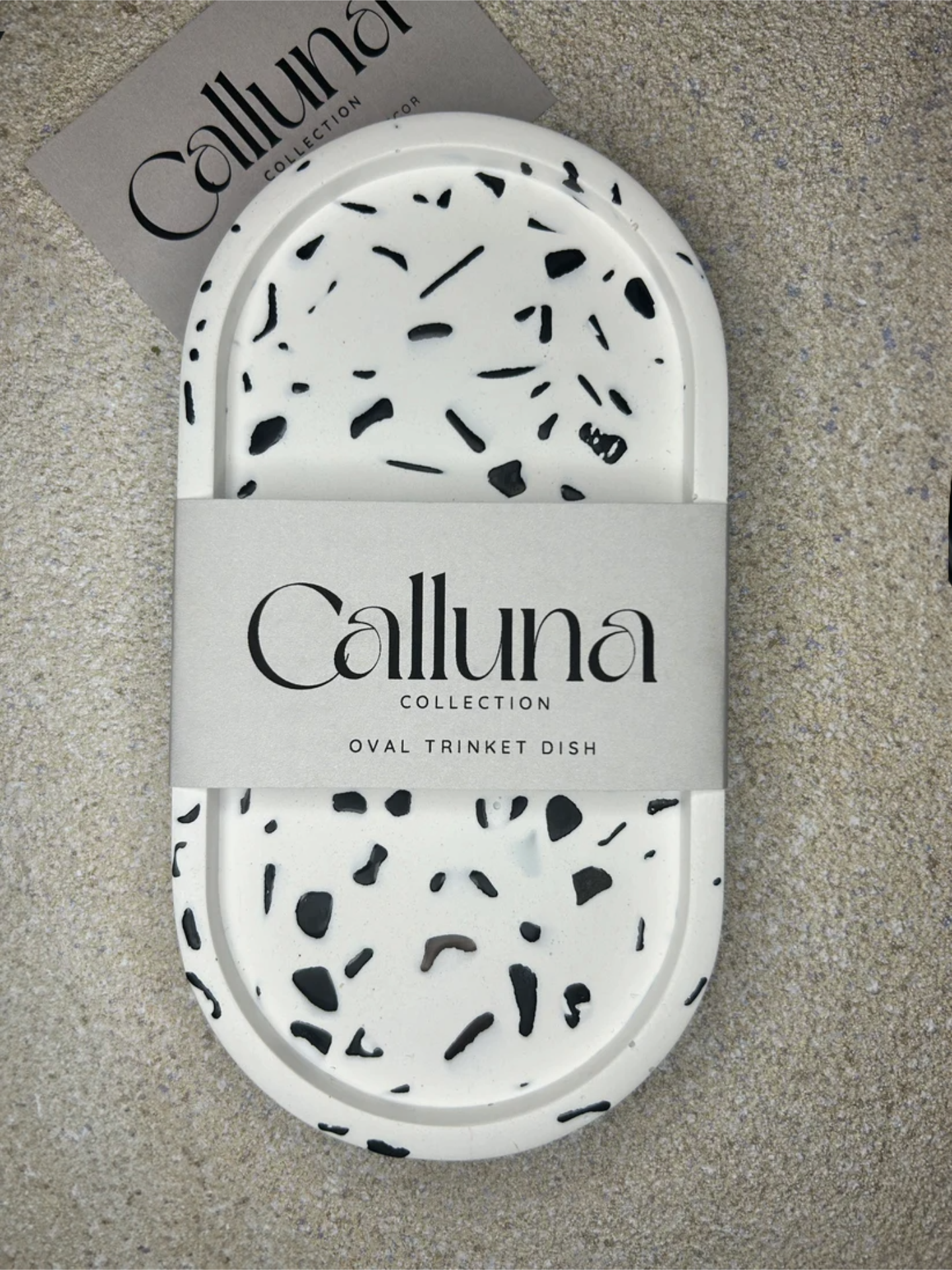 Handmade Home Accessories - A Oval shaped trinket tray that has real black obsidian gemstones in. It has a belly band across the middle saying Calluna Collection which is the brand name. The photo is face on and on a natural stone texture backdrop. It is intended to show a customer how the product is packaged.