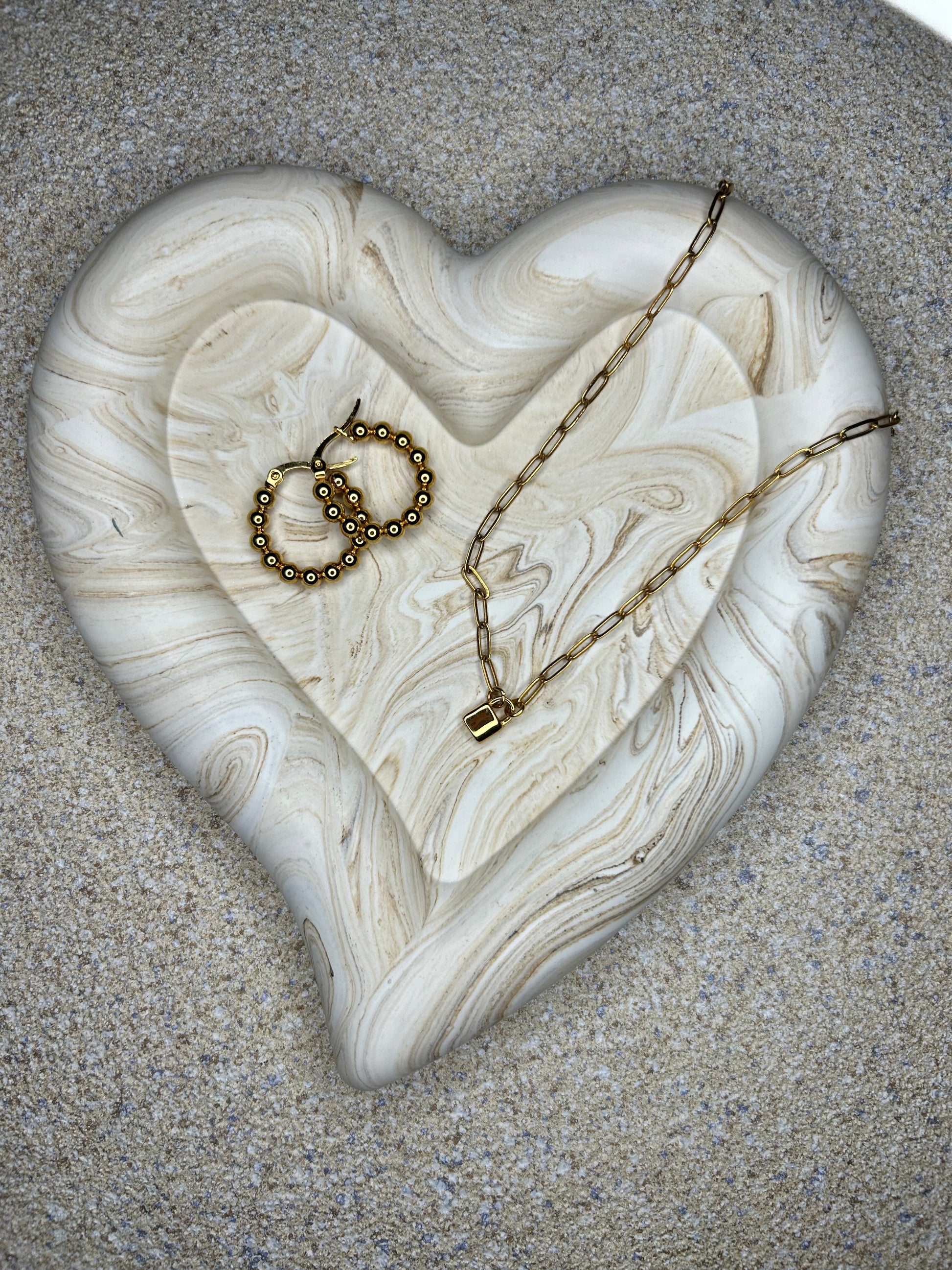 Handmade Home Accessories - A brown marble chunky heart trinket tray, on a natural stone background with a pair of gold hoop earrings and a padlock necklace displayed on it.