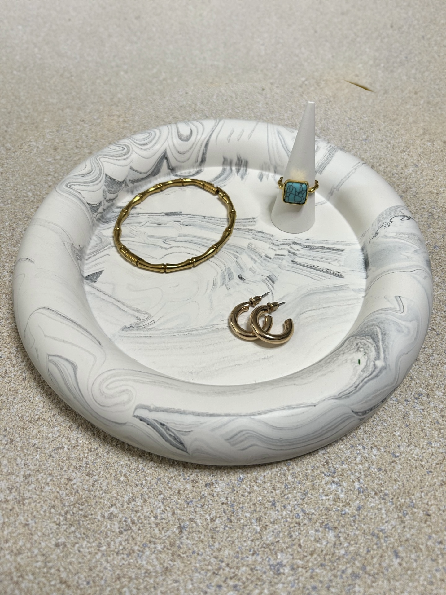 Handmade Home Accessories - A black marbles deep chunky key trinket tray, with a stone textured backdrop. Image taken from the front, highlighting a ring stand with a gold ring on it and a bracelet and hoop earrings.