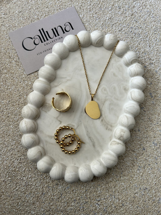 Handmade Home Accessories - A greige marble bubble trinket tray, with a gold ring, necklace and hoop earrings displayed on it. The photo is taken at a 45 degree angle from a birds eye view and is on a stone textured table. In the background is a business card that reads Calluna Collection.