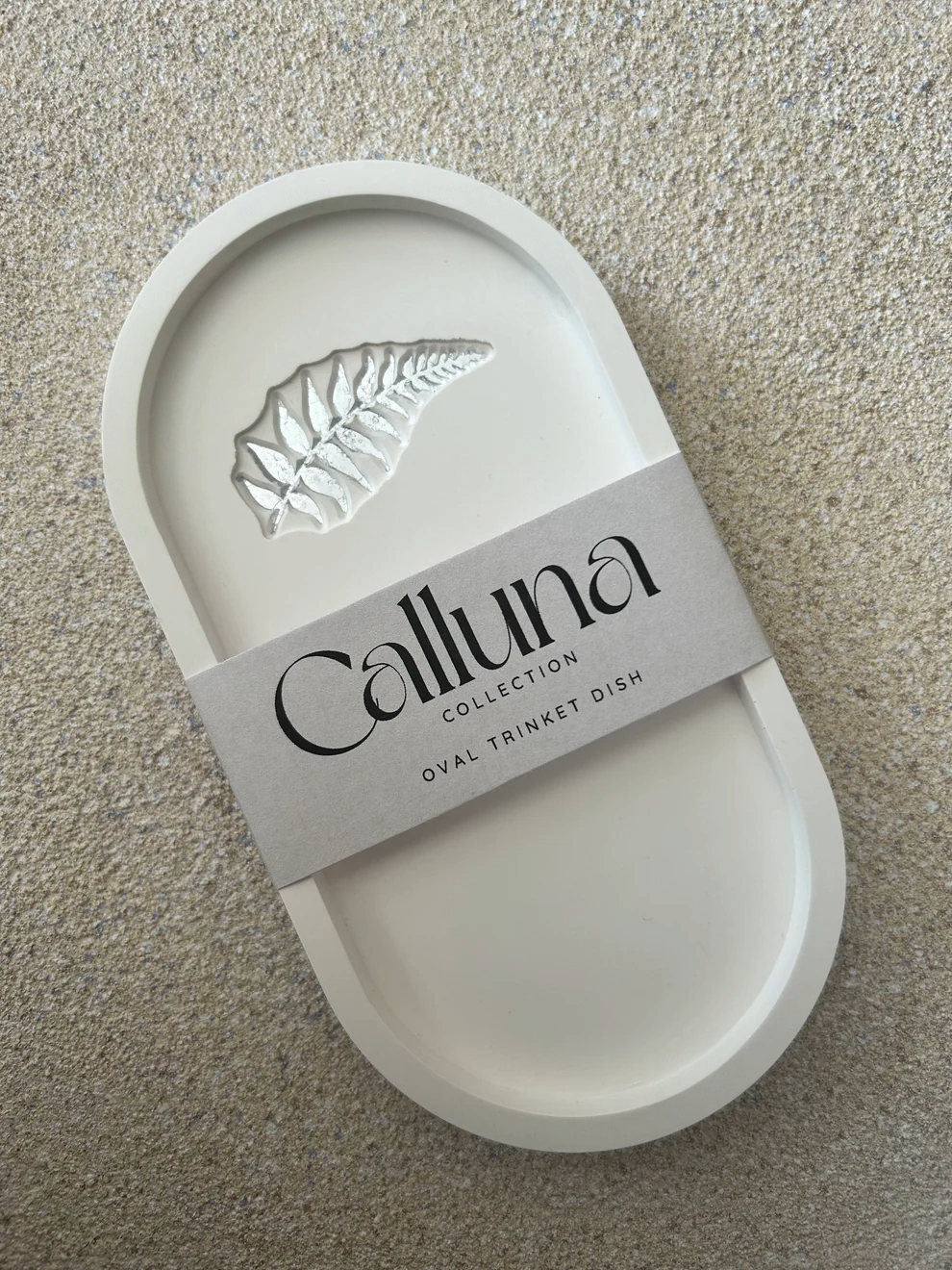 Handmade Home Accessories - A white oval shaped trinket tray with a silver fern leaf detail, it has a belly band around the centre to show its packaging that states the company name Calluna Collection.