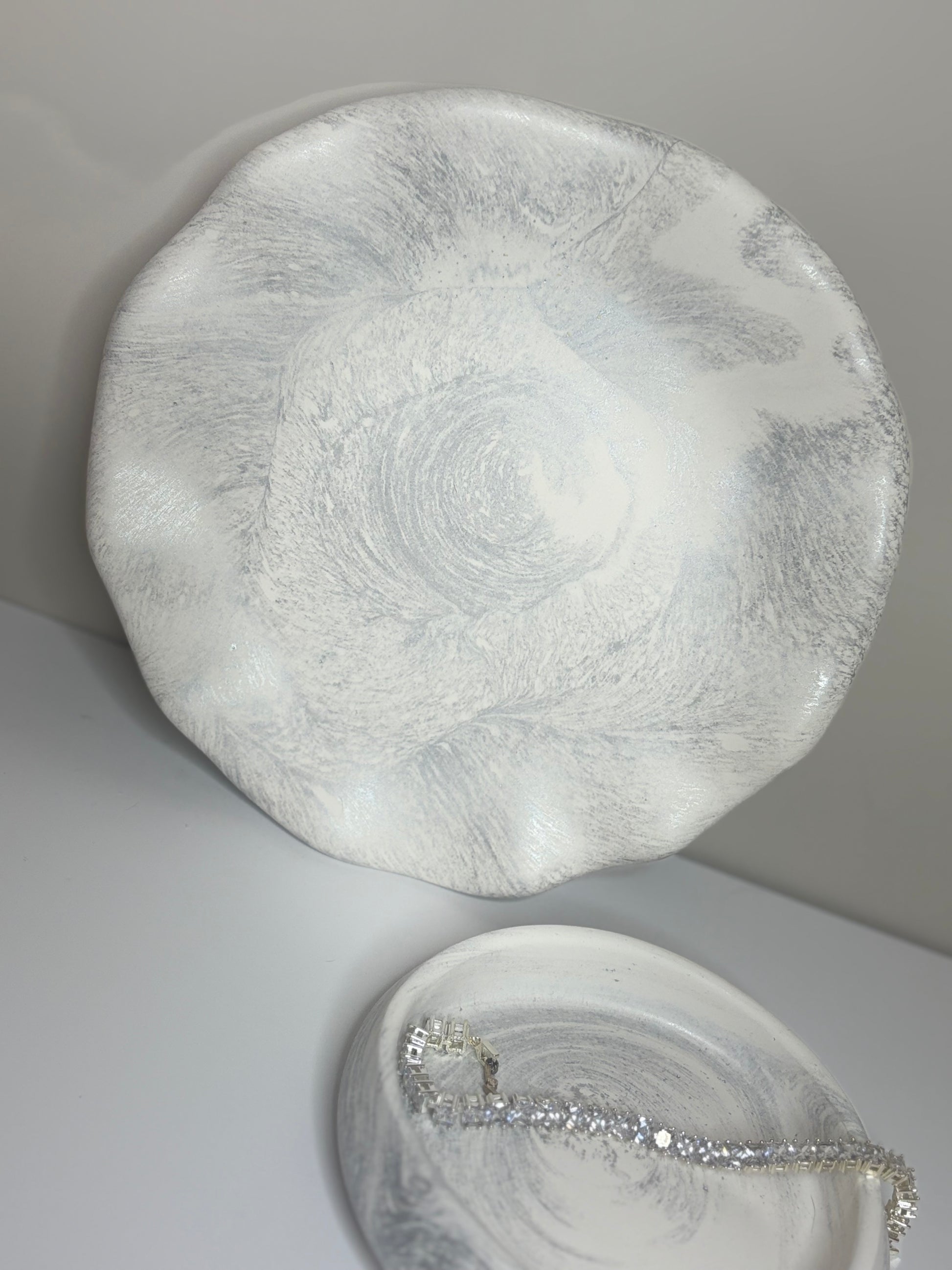 A handmade round decorative plate with a soft grey and white marbled finish, displayed upright beside a smaller matching trinket dish holding a silver bracelet. The plate’s organic, wavy edges and subtle shimmer highlight its elegant, handcrafted design.