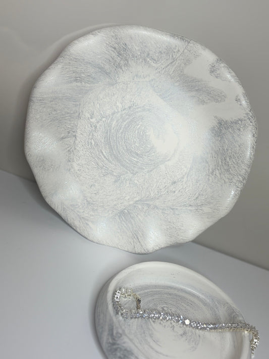 A handmade round decorative plate with a soft grey and white marbled finish, displayed upright beside a smaller matching trinket dish holding a silver bracelet. The plate’s organic, wavy edges and subtle shimmer highlight its elegant, handcrafted design.