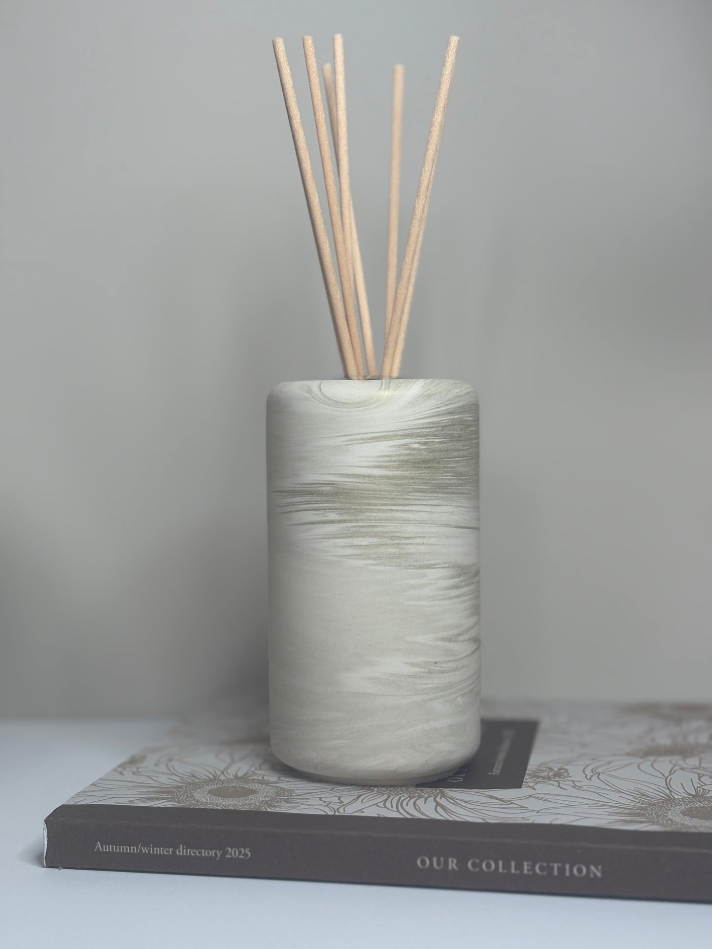 Handmade Solace Diffuser displayed on a neutral-toned book, featuring a tall cylindrical Jesmonite vessel with soft marbled beige and stone grey tones, complemented by natural reed sticks for an elegant minimalist look.