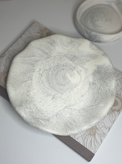 A handmade round decorative plate with a soft grey and white marbled design, crafted from Jesmonite. The plate rests on a floral-patterned book, showcasing its smooth matte texture and organic swirling detail.