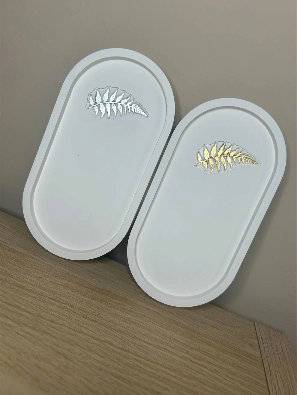 Handmade Home Accessories - twowhite oval trinket trays with fern leaf details, one in gold and one in silver. They are propped against a light brown wall and on a light oak base.