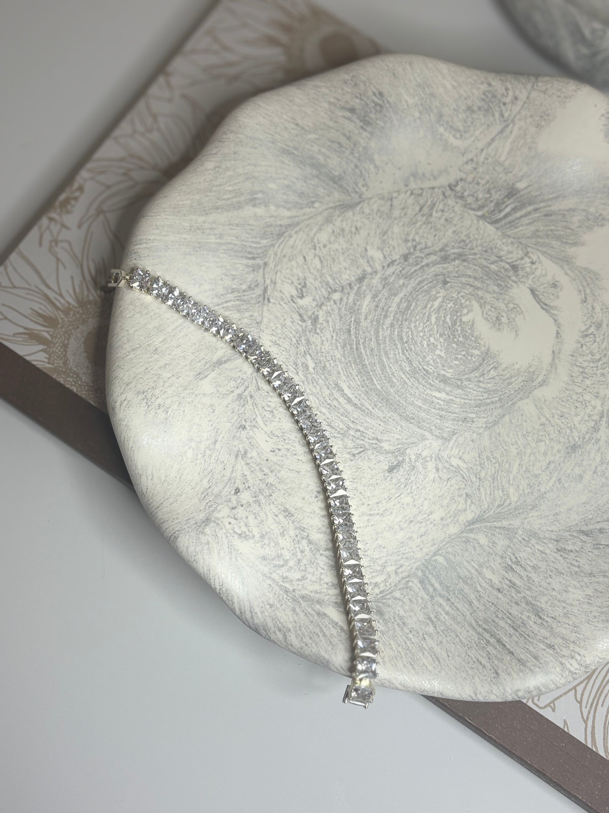 A handmade round decorative plate with a soft grey and white marbled design, featuring a silver tennis bracelet laid elegantly across its surface. The plate rests on a floral-patterned book, showcasing its smooth matte texture and wavy organic edge.