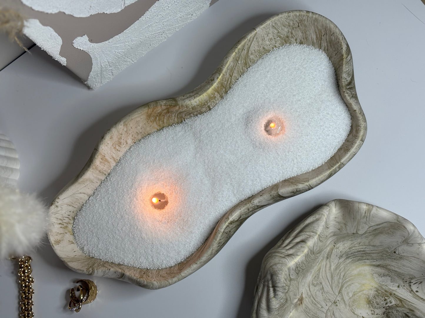 A handcrafted Jesmonite tray with soft, organic curves and a neutral marbled finish, filled with white sand and two softly glowing tealight candles. Styled on a minimal surface alongside gold jewellery, the Elysian Glow Tray exudes a calming, luxurious aesthetic.