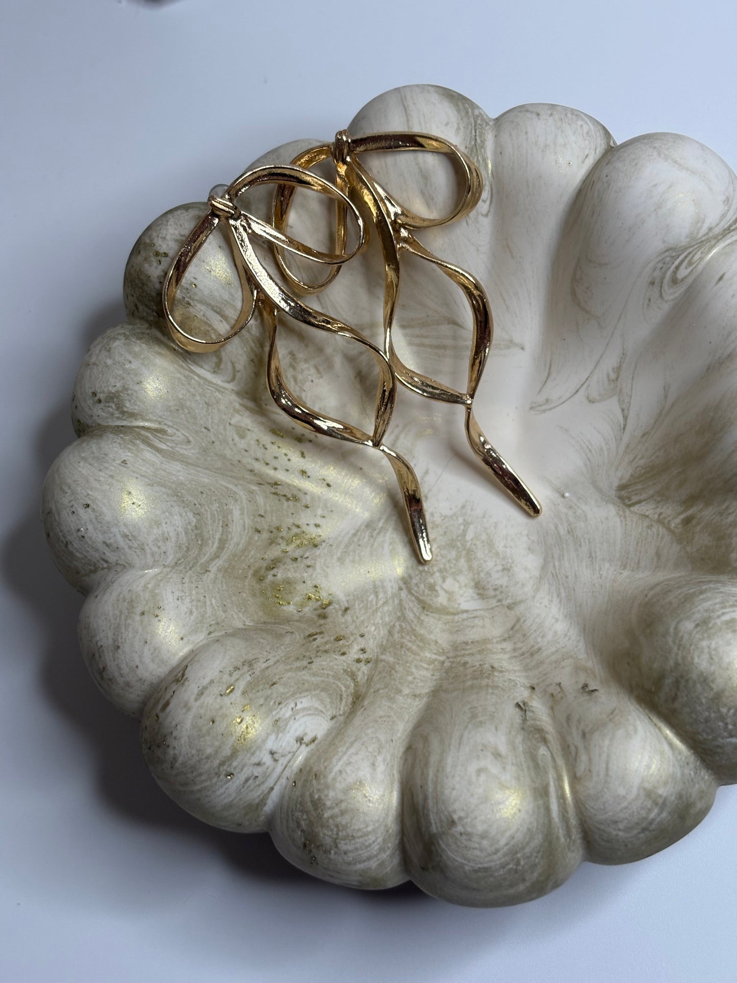A handmade Jesmonite Celeste Trinket Bowl with a sculptural, rippled edge and soft gold marbled detailing, displayed with a pair of elegant gold bow-shaped earrings resting inside. The bowl’s organic form and muted tones create a refined, minimalist aesthetic.