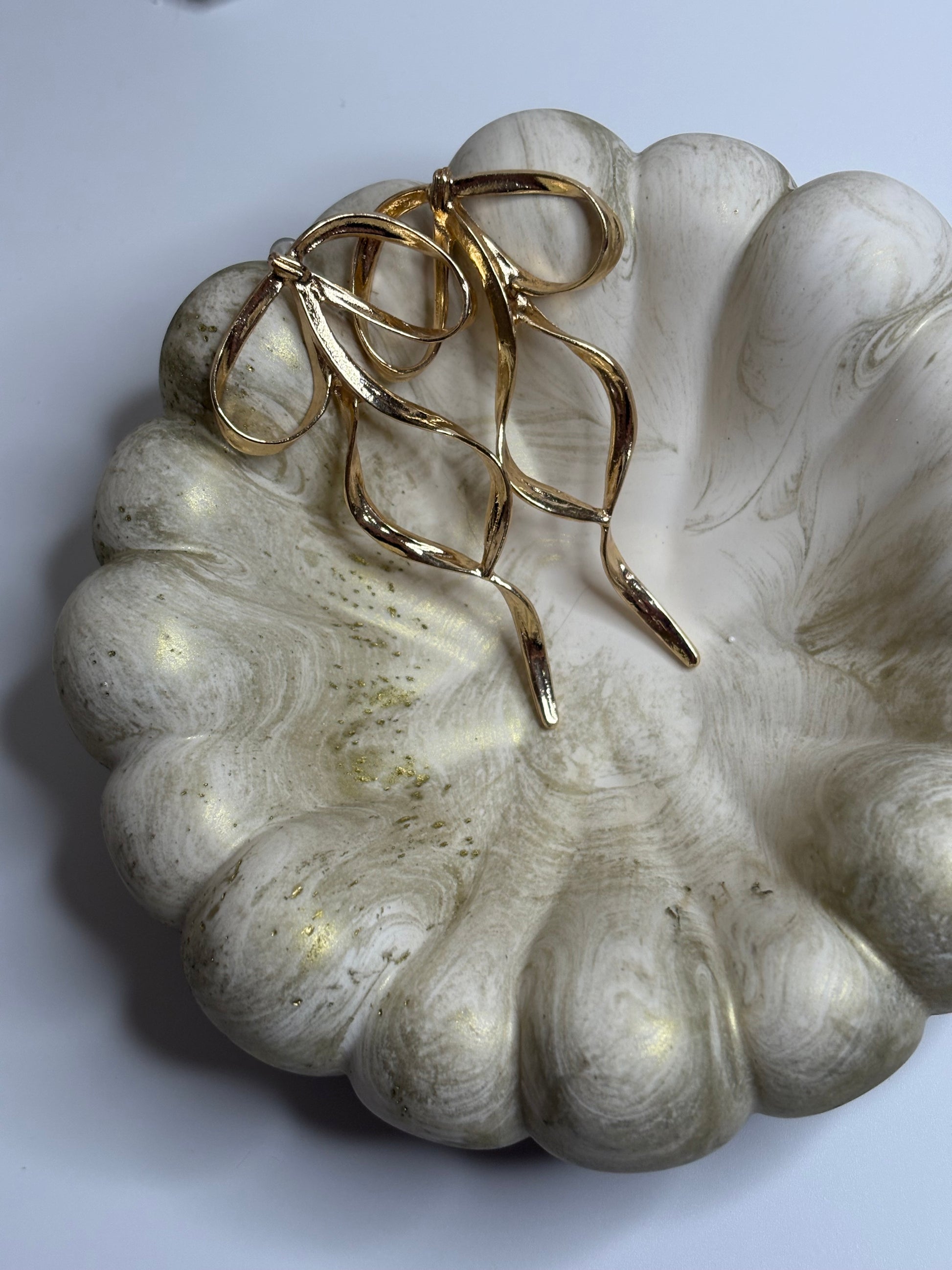 A handmade Jesmonite Celeste Trinket Bowl with a sculptural, rippled edge and soft gold marbled detailing, displayed with a pair of elegant gold bow-shaped earrings resting inside. The bowl’s organic form and muted tones create a refined, minimalist aesthetic.