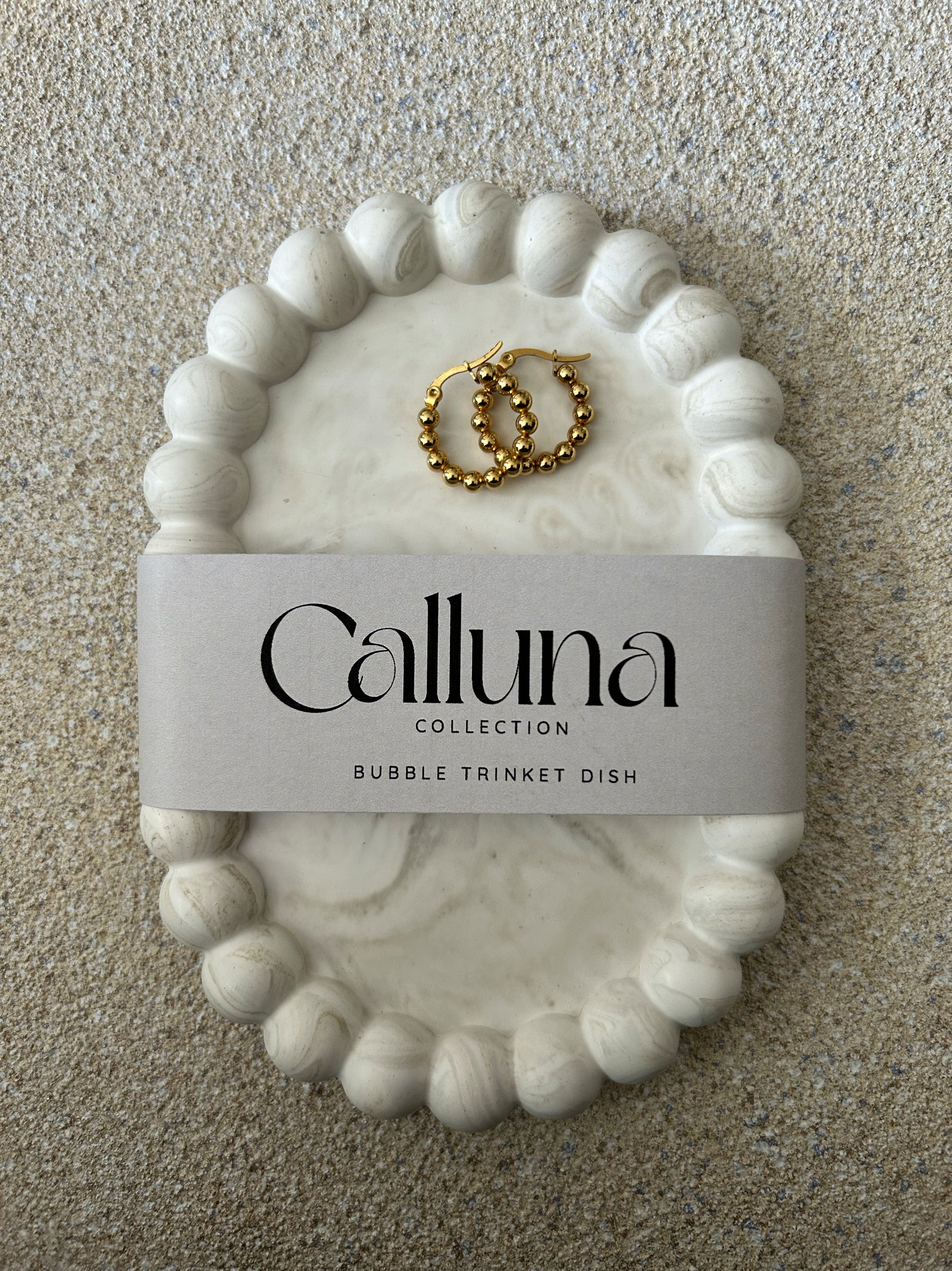 Handmade Home Accessories - A Greige marble bubble tray, with a pair of bubble style gold hoop earrings on. The photo is taken from a birds eye view and displays a belly band with the company name Calluna Collection. The photo is taken to provide a visual of what the product looks like packaged.