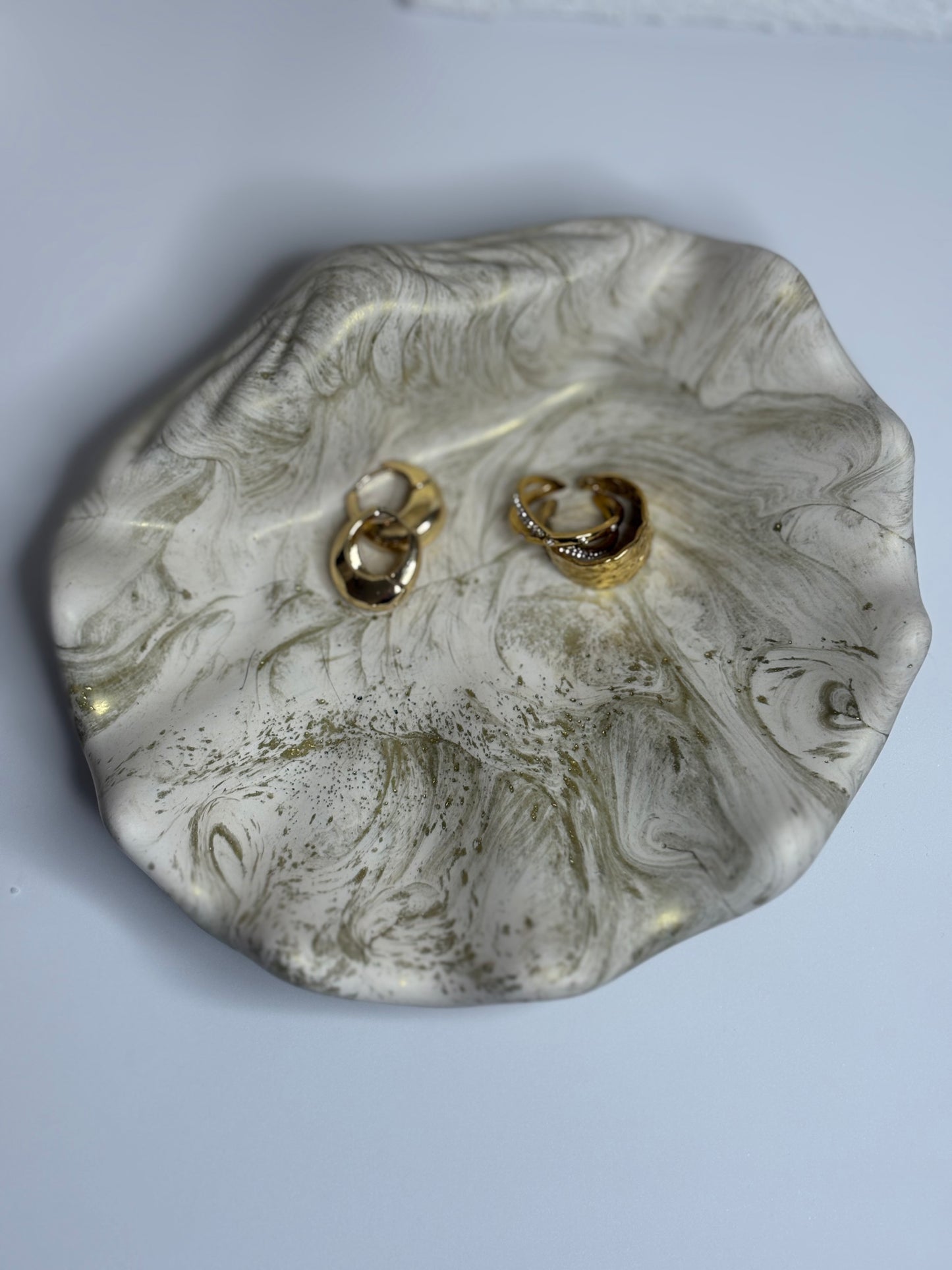 A handcrafted Jesmonite trinket dish shaped like a soft shell, featuring swirling marbled tones of ivory and muted gold. The dish holds two gold rings and rests on a clean white surface, showcasing its organic texture and elegant, sculptural design.