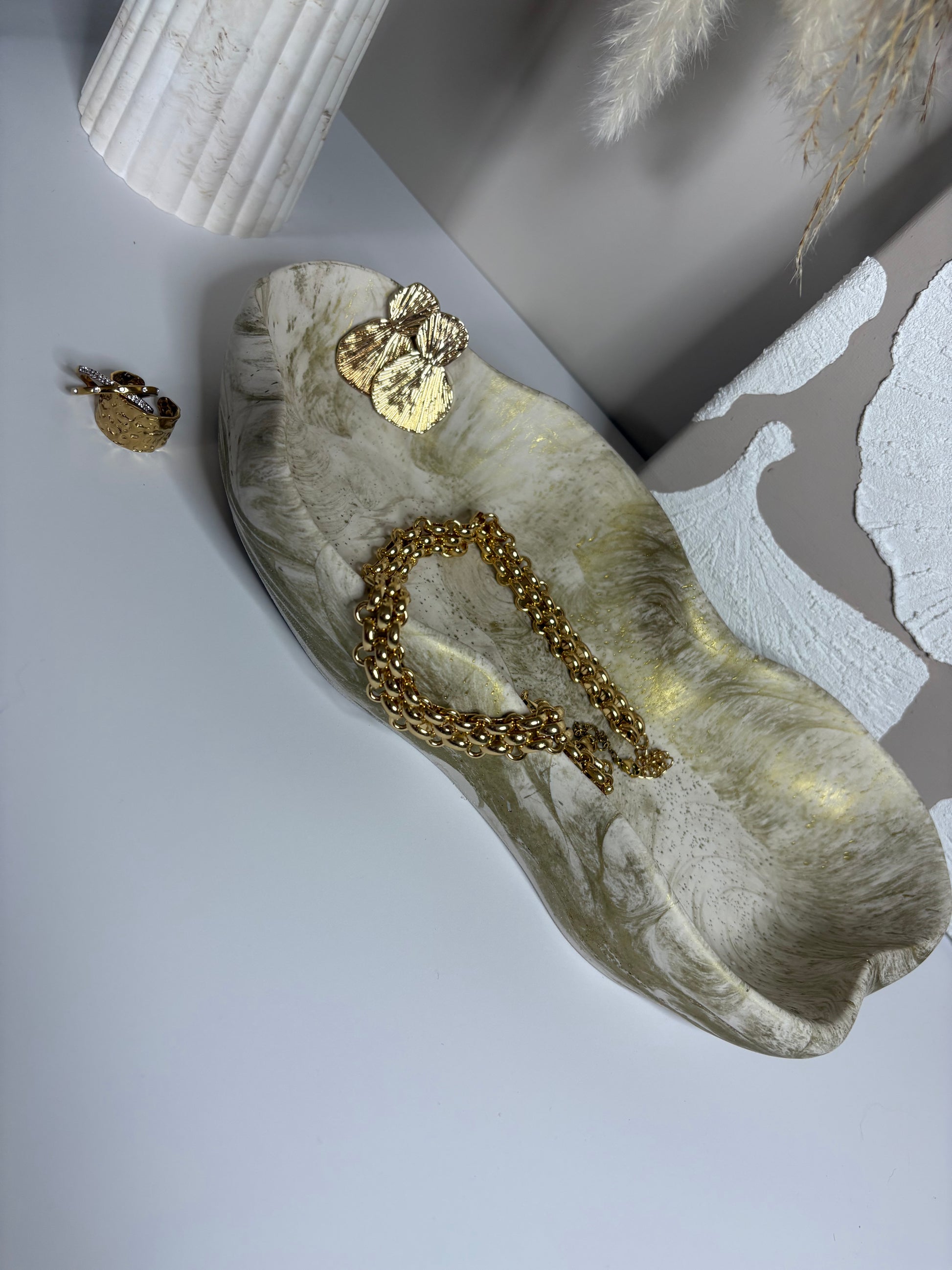 A handcrafted Jesmonite tray with soft, organic curves and a marbled neutral finish, styled with gold jewellery including a chunky chain and shell-shaped earrings. Set beside a ribbed vase and textured wall art, the Elysian Glow Tray adds understated elegance to a modern interior.