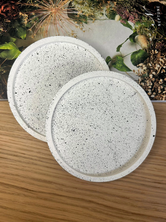 Set of two round white Jesmonite coasters with black splatter detailing, styled with floral textures and natural wood – minimalist handmade accessories.
