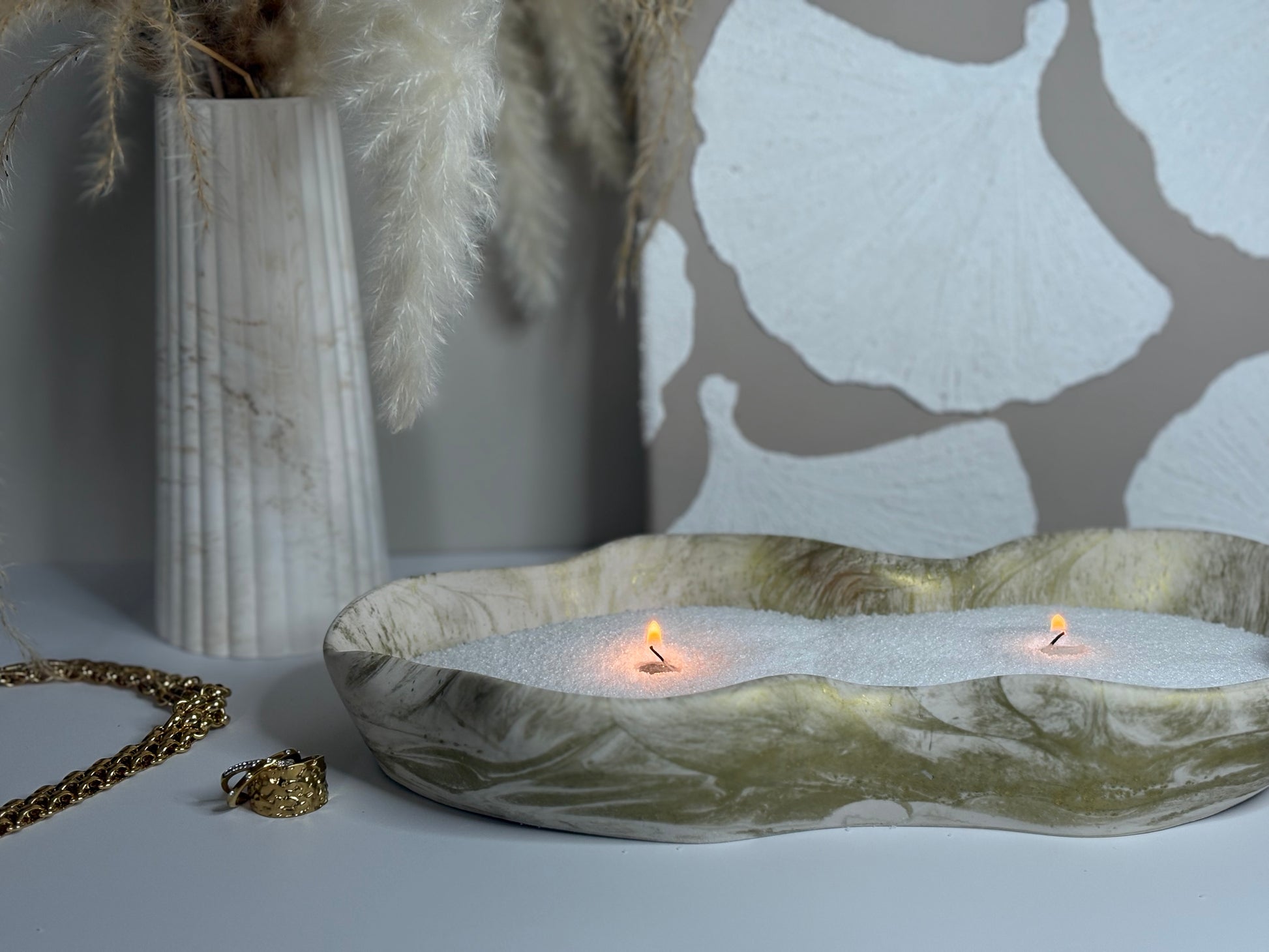 A handcrafted Jesmonite tray with organic curves and a marbled neutral finish, filled with white sand and two softly glowing tealight candles. Styled beside a ribbed ceramic vase of pampas grass and gold jewellery, the Elysian Glow Tray creates a serene, minimalist ambience.