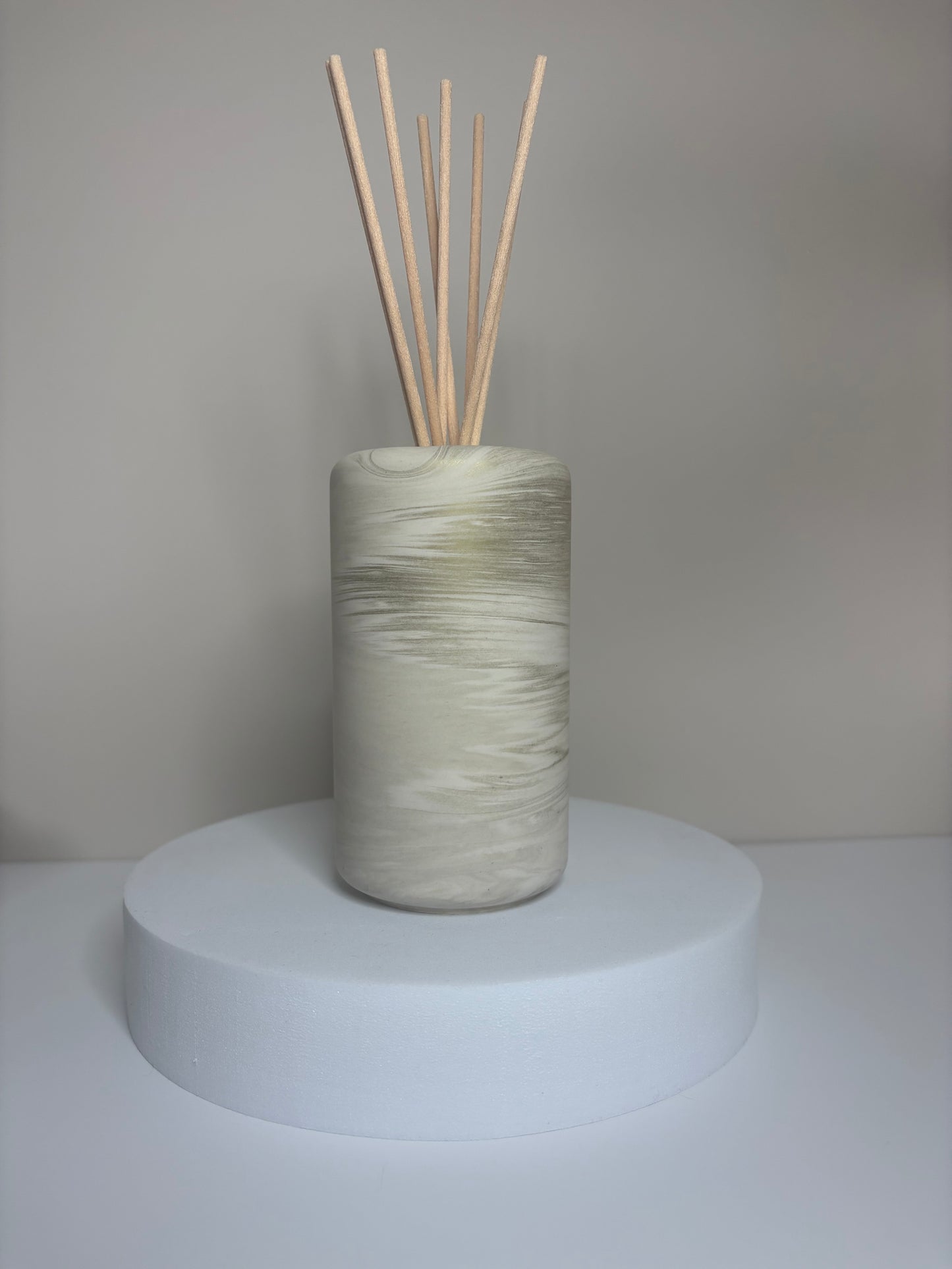 Handmade Solace Diffuser featuring a tall cylindrical Jesmonite vessel with soft marbled tones of beige and stone grey, displayed on a round white pedestal with natural reed sticks.