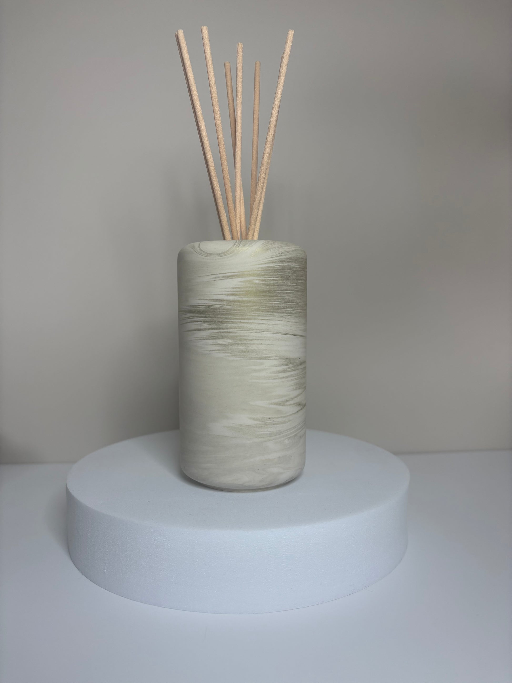 Handmade Solace Diffuser featuring a tall cylindrical Jesmonite vessel with soft marbled tones of beige and stone grey, displayed on a round white pedestal with natural reed sticks.