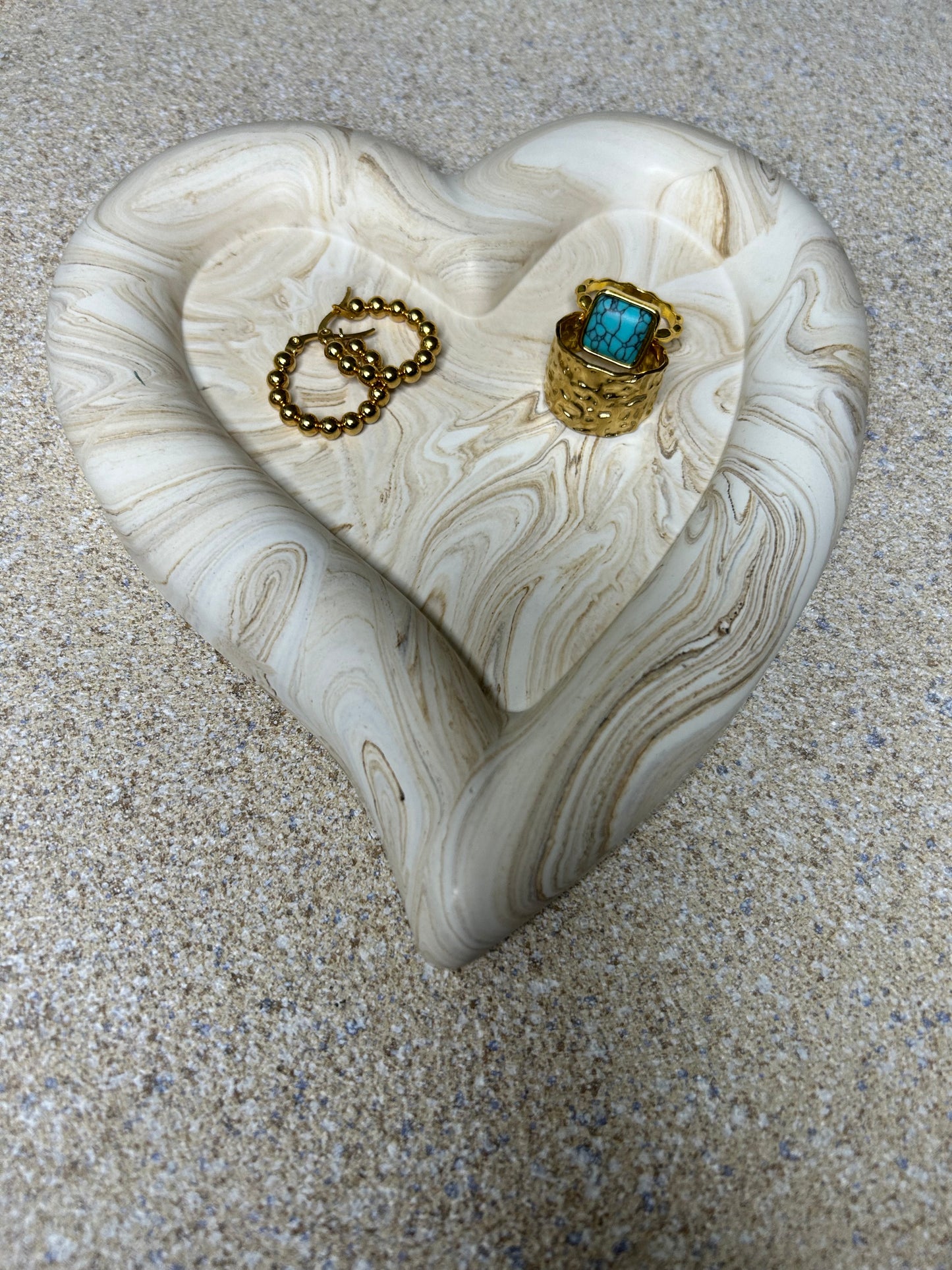 Handmade Home Accessories - A angled shot of a heart shaped trinket tray that is brown marble effect with 2 gold rings on and a pair of gold hoops. based on a natural stone background.