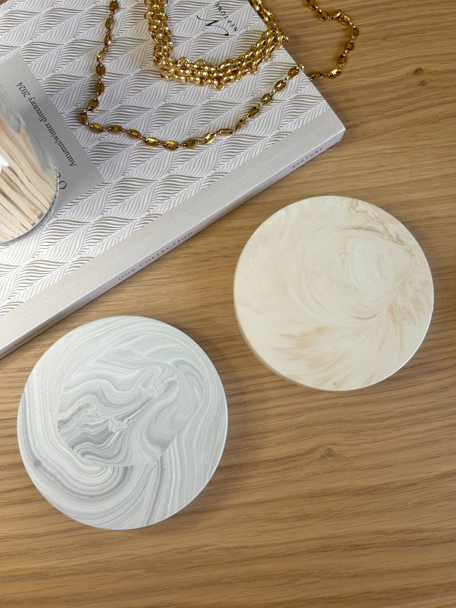 Two handcrafted Jesmonite coasters in swirling grey and beige tones, styled with gold jewellery – minimalist round home accessories.
