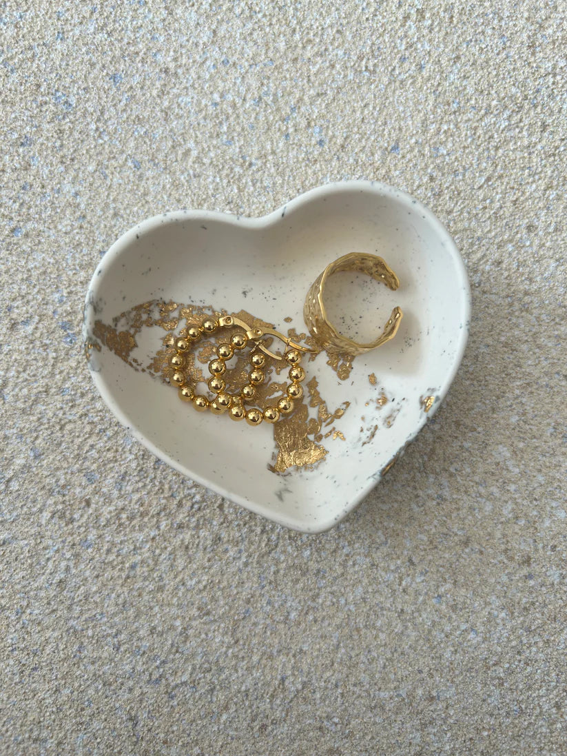 Handmade Home Accessories - A small heart shaped trinket dish, that is a cream colour with black splatter and gold leaf details. The dish is holding a pair of gold hoop earrings and a gold ring.