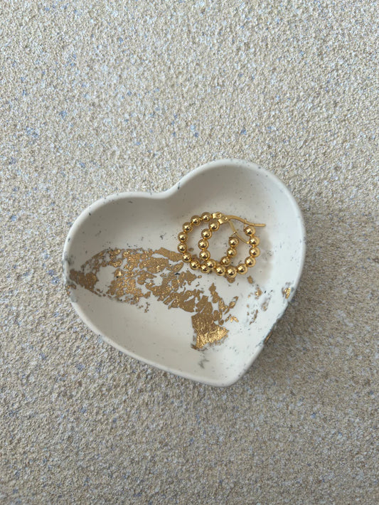 Handmade Home Accessories - A small heart shaped trinket dish, that is a cream colour with black splatter and gold leaf details. The dish is holding a pair of gold hoop earrings.