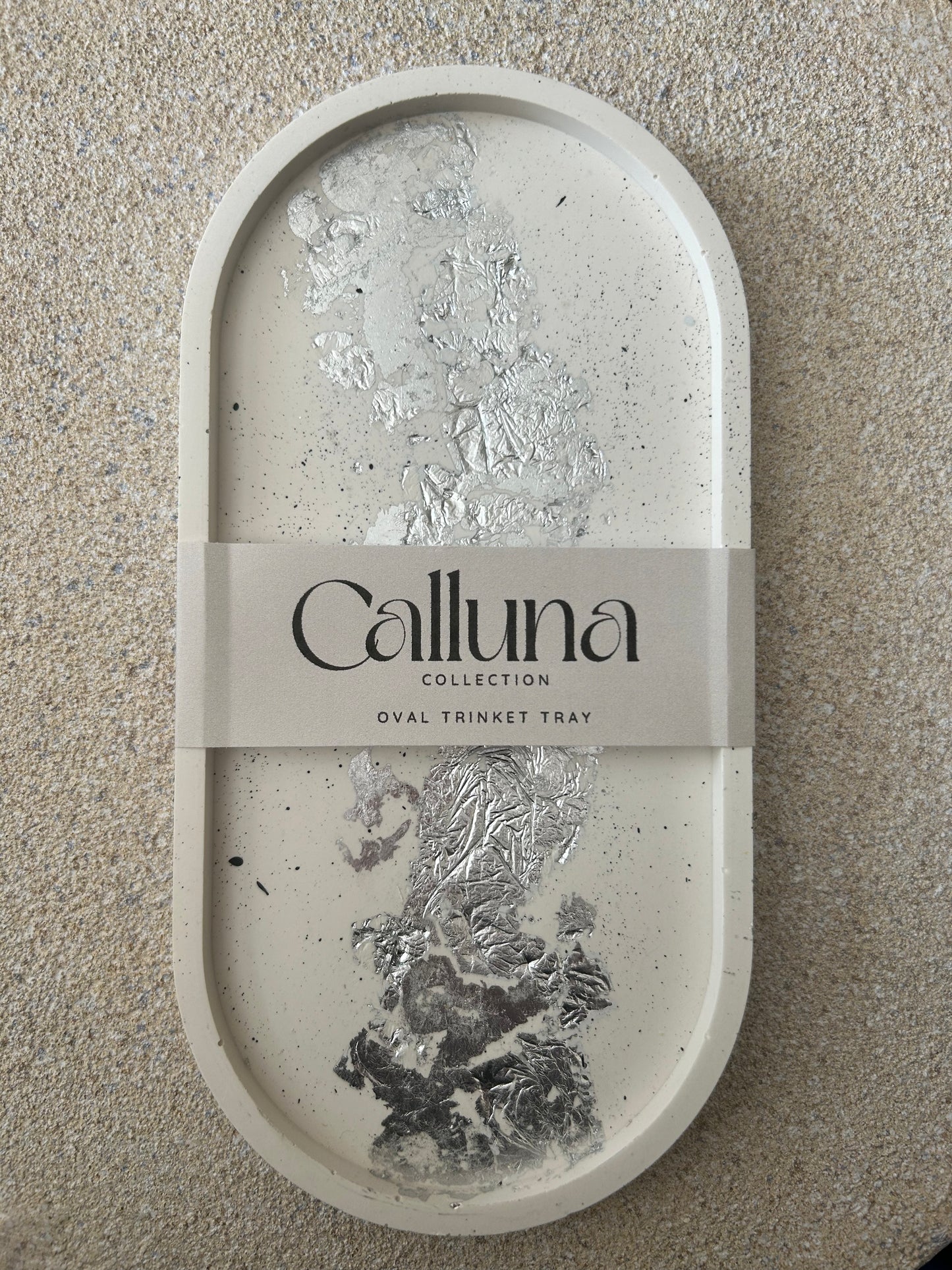 Handmade Home Accessories - A Large oval cream trinket tray with black splatters and silver leaf detail. It has a belly band around the centre sating Calluna Collection which is the brand.