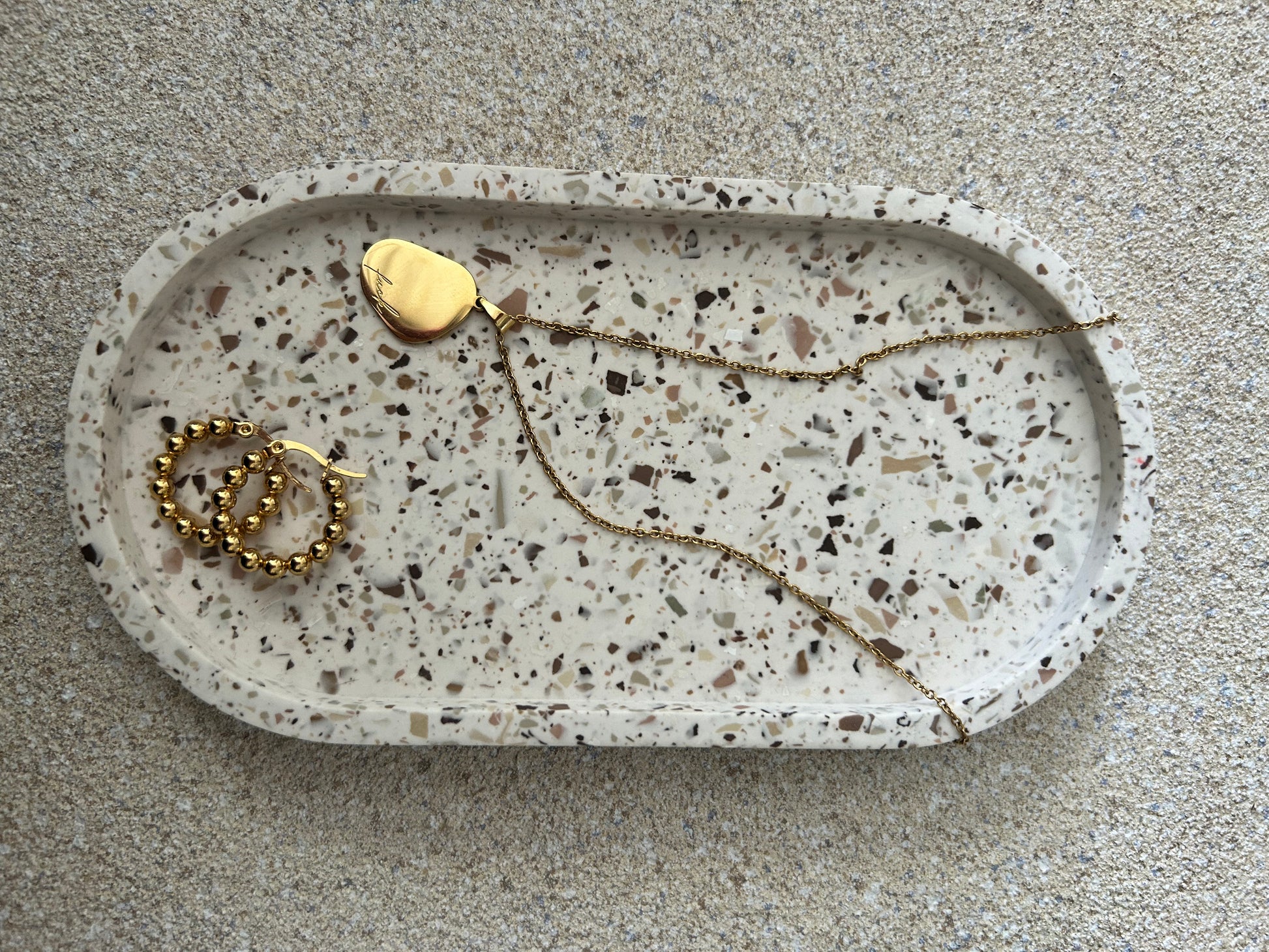 Handmade Home Accessories - A single terrazzo oval trinket tray in  an off white colour which has been styled with a pair of gold hoops and a gold necklace.