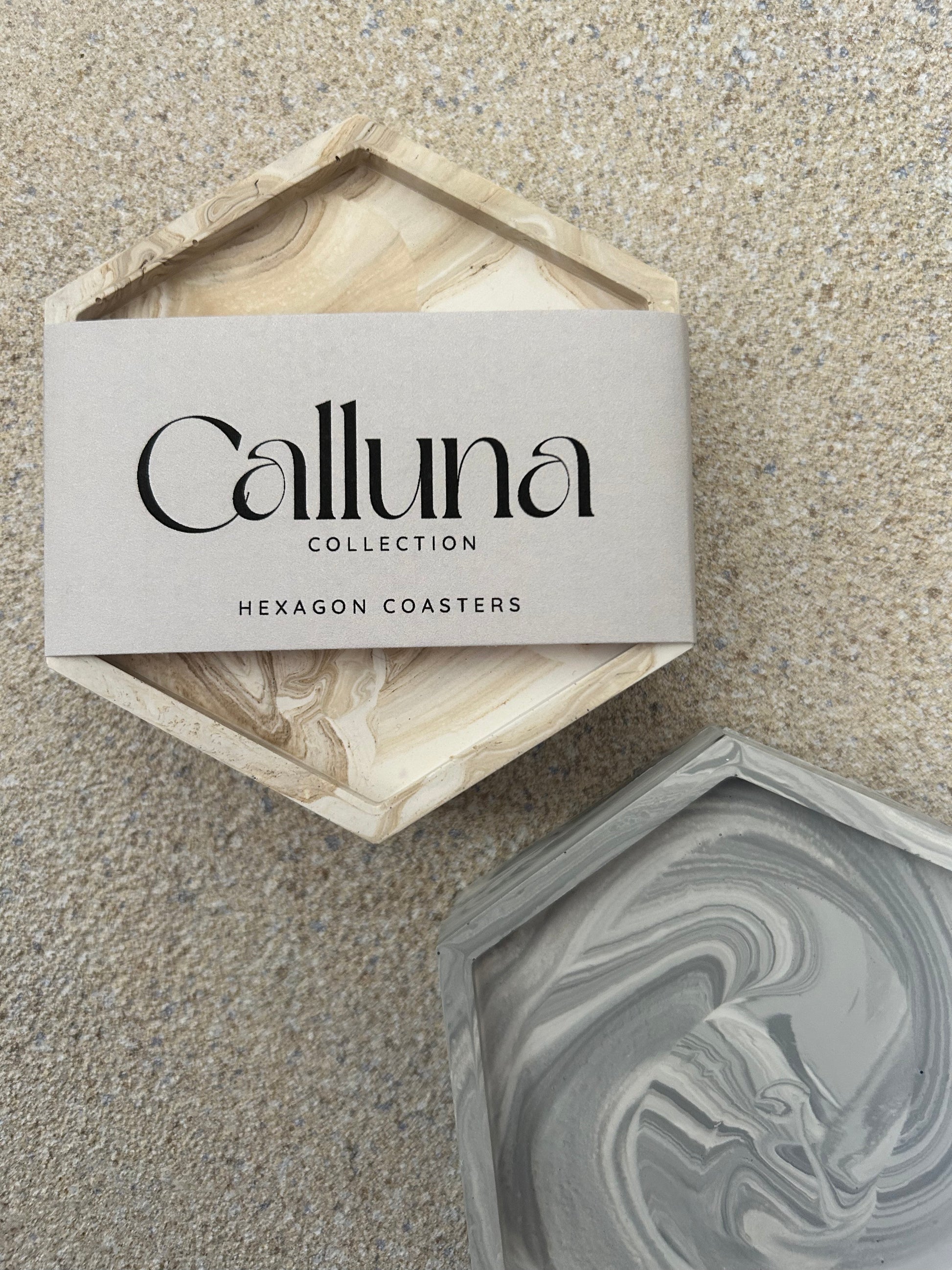 Handmade Home Accessories - A beigemarble and grey marble hexagon coaster shot from birds eye view, the beige coaster has a belly band around it that states the brand name Calluna Collection.