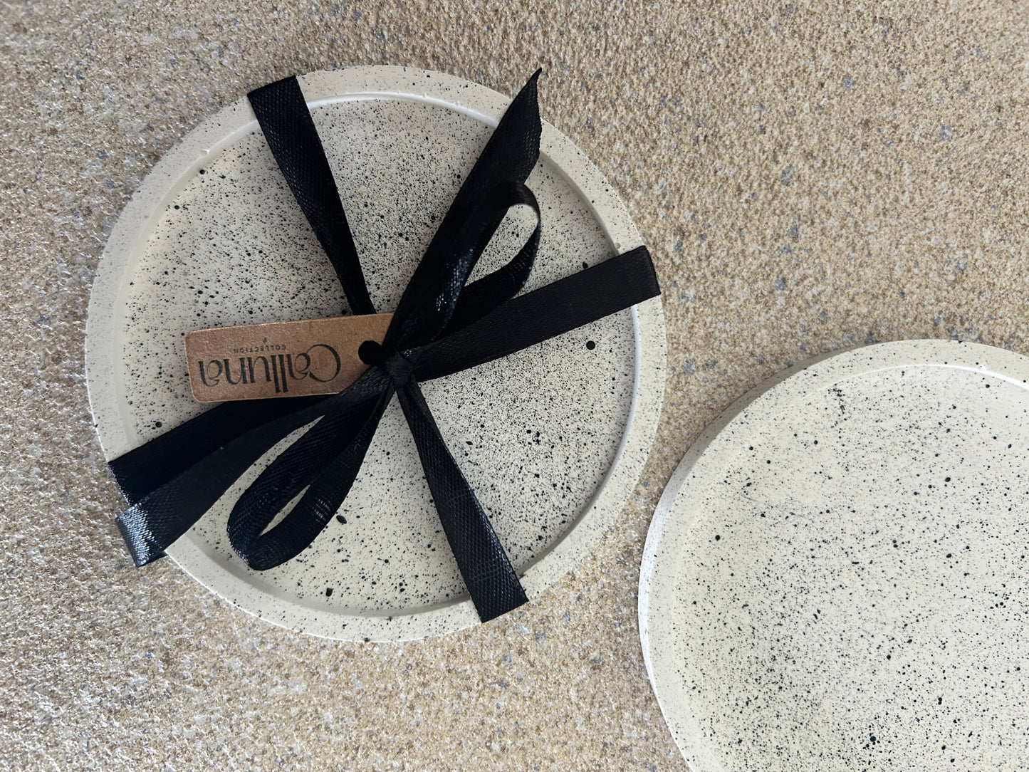 Handmade Home Decor - A picture of a stack of white coasters with black splatters, covered with a black tied ribbon with a tag on saying Calluna Collection.