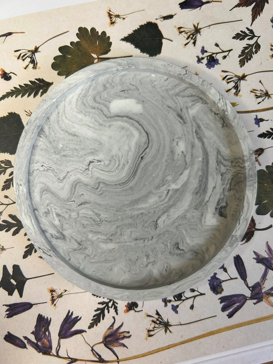 Close-up of handmade round Jesmonite coaster in swirling grey marble pattern, styled on a floral book cover – refined decorative home accessory.