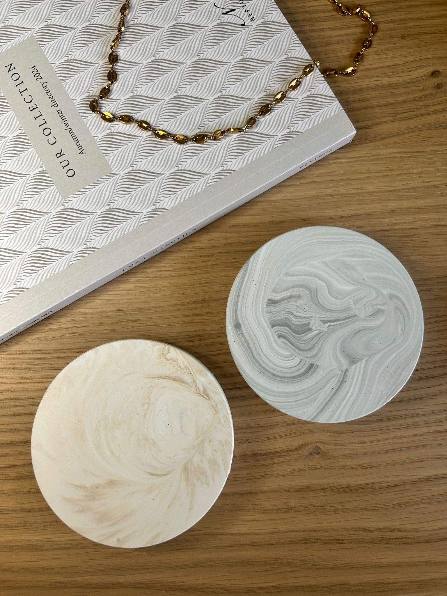 Pair of round Jesmonite coasters in beige and grey marbled finishes, styled on a wooden surface with jewellery and textured paper.