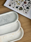 Set of three handmade Jesmonite trays in grey, ivory, and warm cream marble finishes, styled beside a botanical book – sustainable and versatile home accessories.