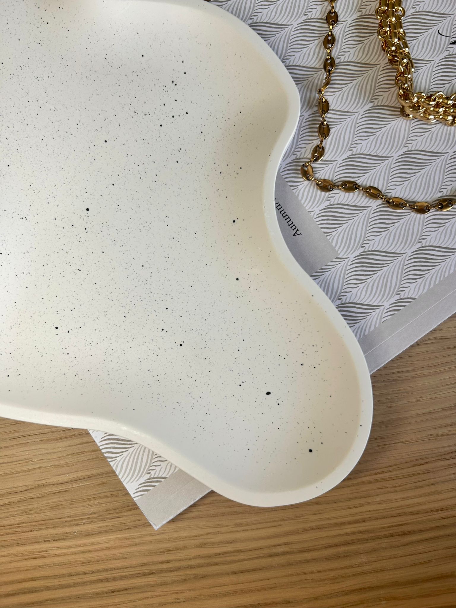 Close-up of organic-shaped white Jesmonite tray with subtle black speckle pattern, styled beside gold chain jewellery – modern handmade home accessory.