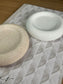 Two round Jesmonite trays with natural speckled textures in beige and white, displayed on a patterned book. A sculptural set of handmade home accessories.
