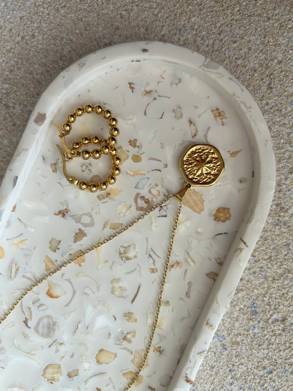 Handmade Home Accessories - A close up shot of an oval trinket tray that has real seashells in a terrazzo design, it has a pair of gold hoop earrings and a star coin style necklace on to display.