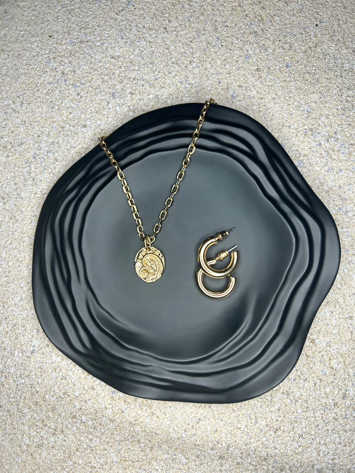 Handmade Home Accessories - Black Ripple trinket dish, the photo is taken from a birds eye view. It has a gold necklace and a gold pair of hoops displayed on it and the background is a textured natural stone.