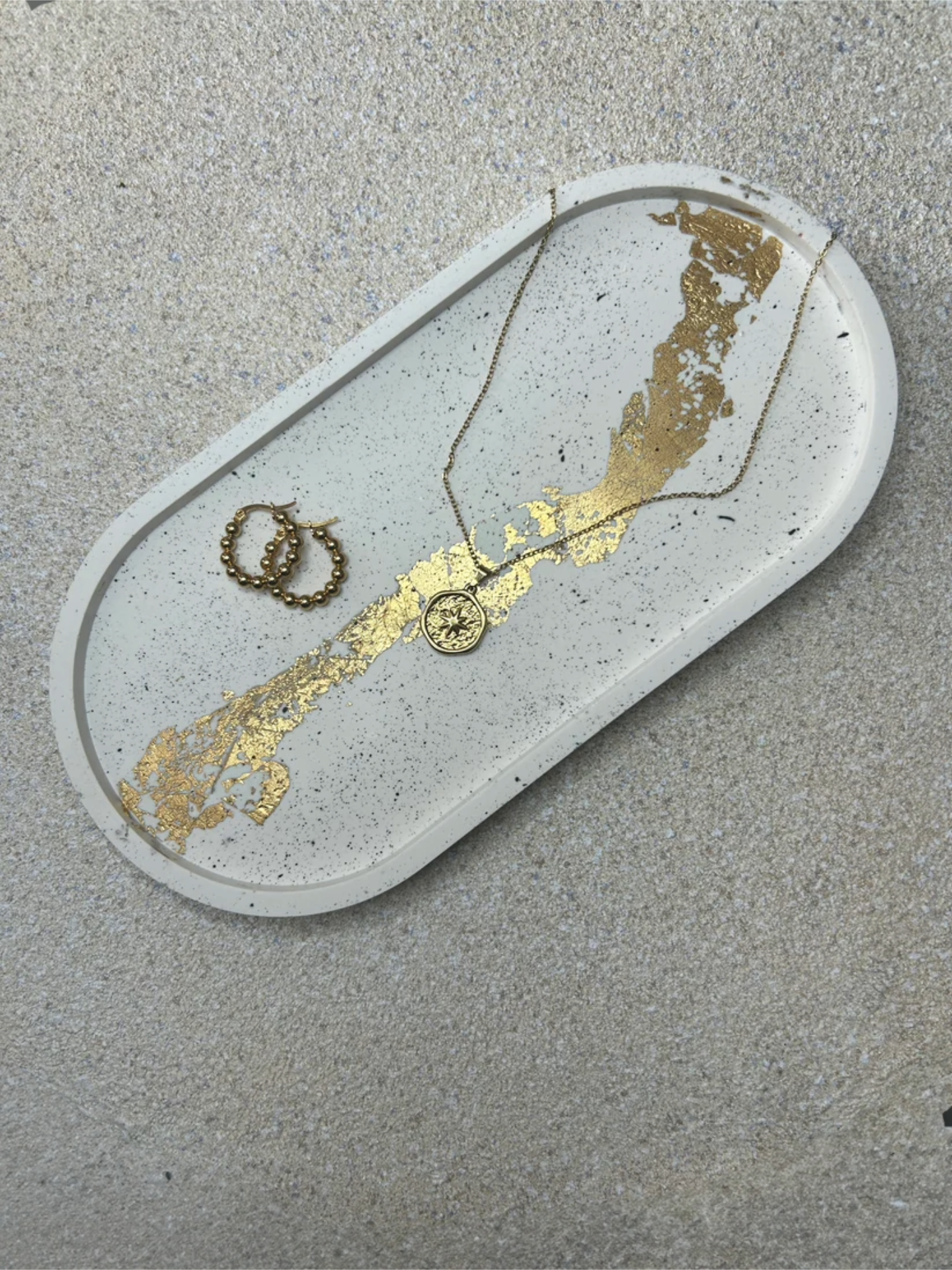 Handmade Home Accessories - Large Oval Trinket tray on a natural stone background, with cream main bod, that has black splatters and gold leaf detail. The picture is a 45 degree angle and the tray has a pair of gold hoop earrings and a star style coin necklace on for decorative purposes.