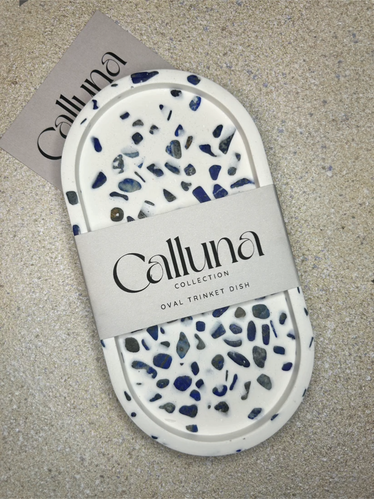 Handmade Home Accessories - Blue and white oval shaped trinket tray made from lapis lazuli gemstones on a background of natural stone. The photo of front on and it has a belly band on with the company logo, and the words Calluna Collection. It is intended to show the customer how the product is packaged.